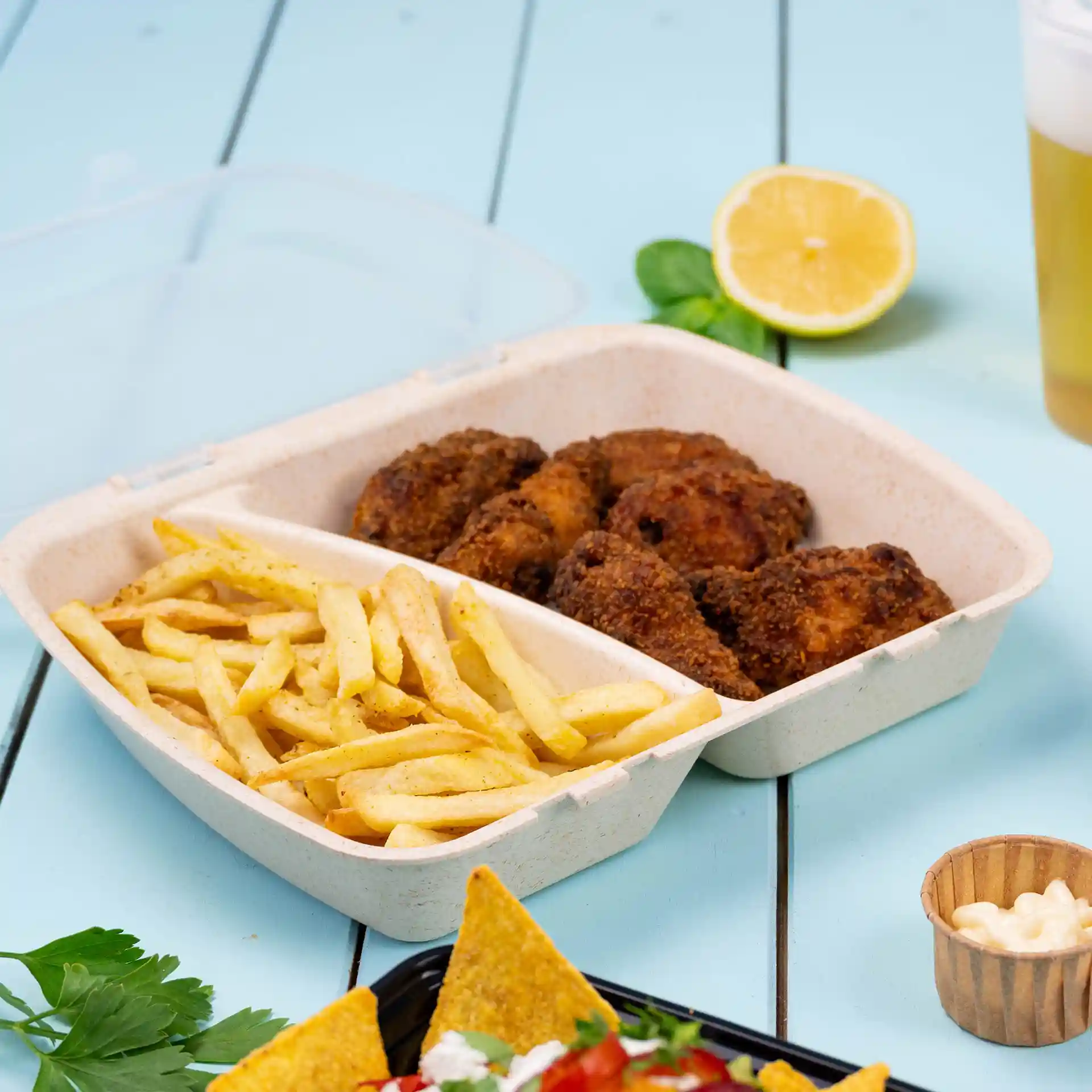 Container met twee compartimenten met gefrituurde kip en frietjes, op een blauwe ondergrond met een citroen en een drankje erbij.