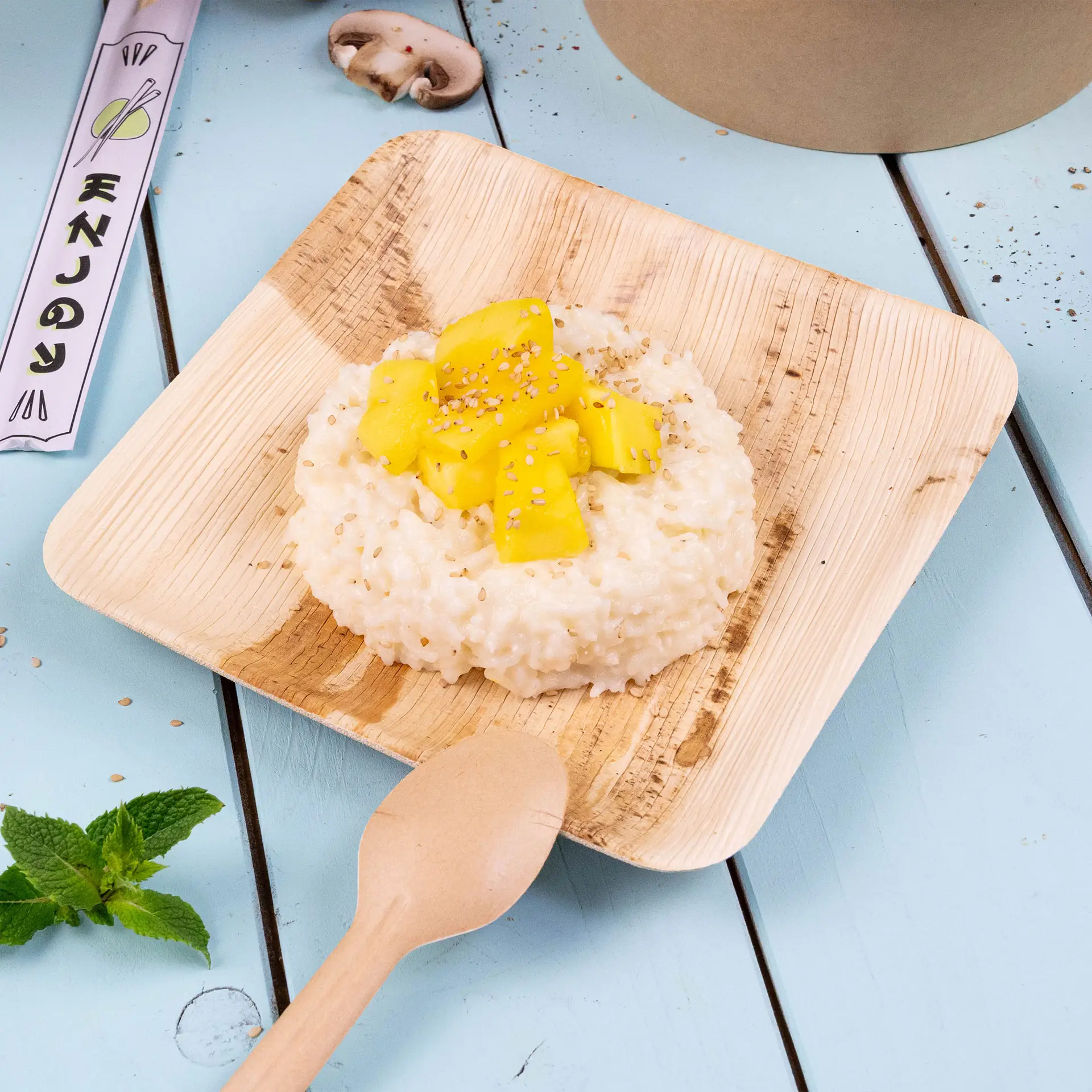 Vierkant palmblad bord met risotto, getopt met mango en sesamzaadjes, vergezeld van een houten lepel en muntblaadjes.