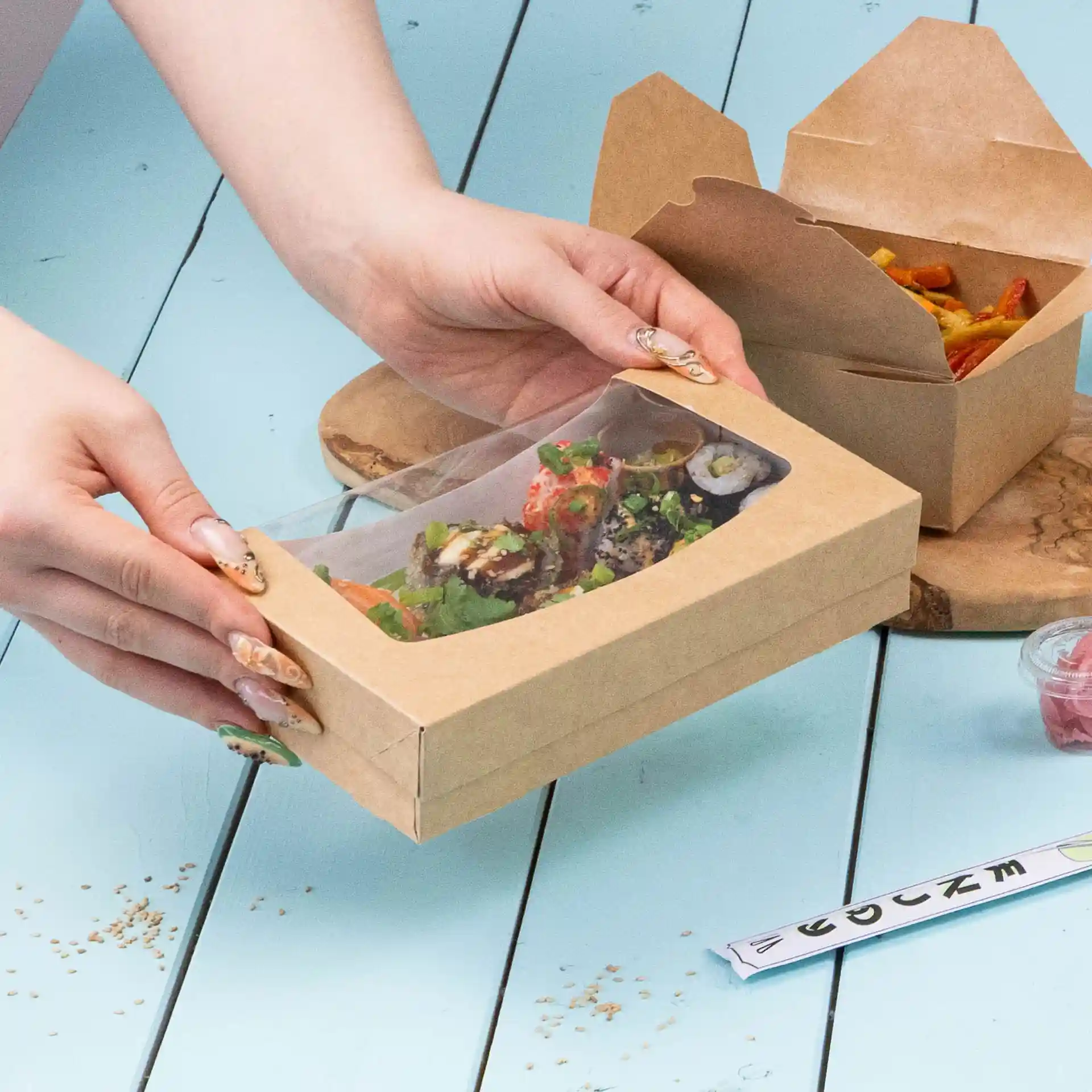 Kartonvensterdoos met sushi en salade, met een transparante bovenkant, op een blauwe ondergrond.