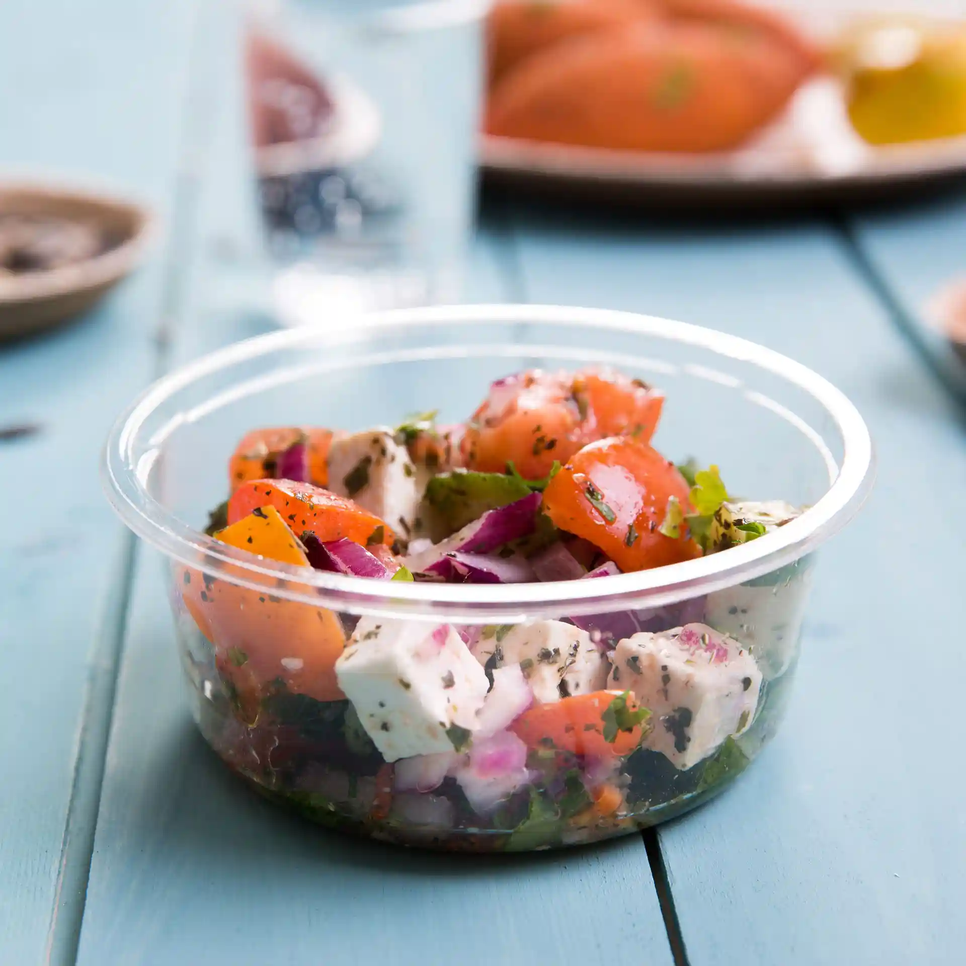 Transparante ronde deli-container met een salade-mix van tomaten, uien en kruiden, geplaatst op een blauwe houten ondergrond.