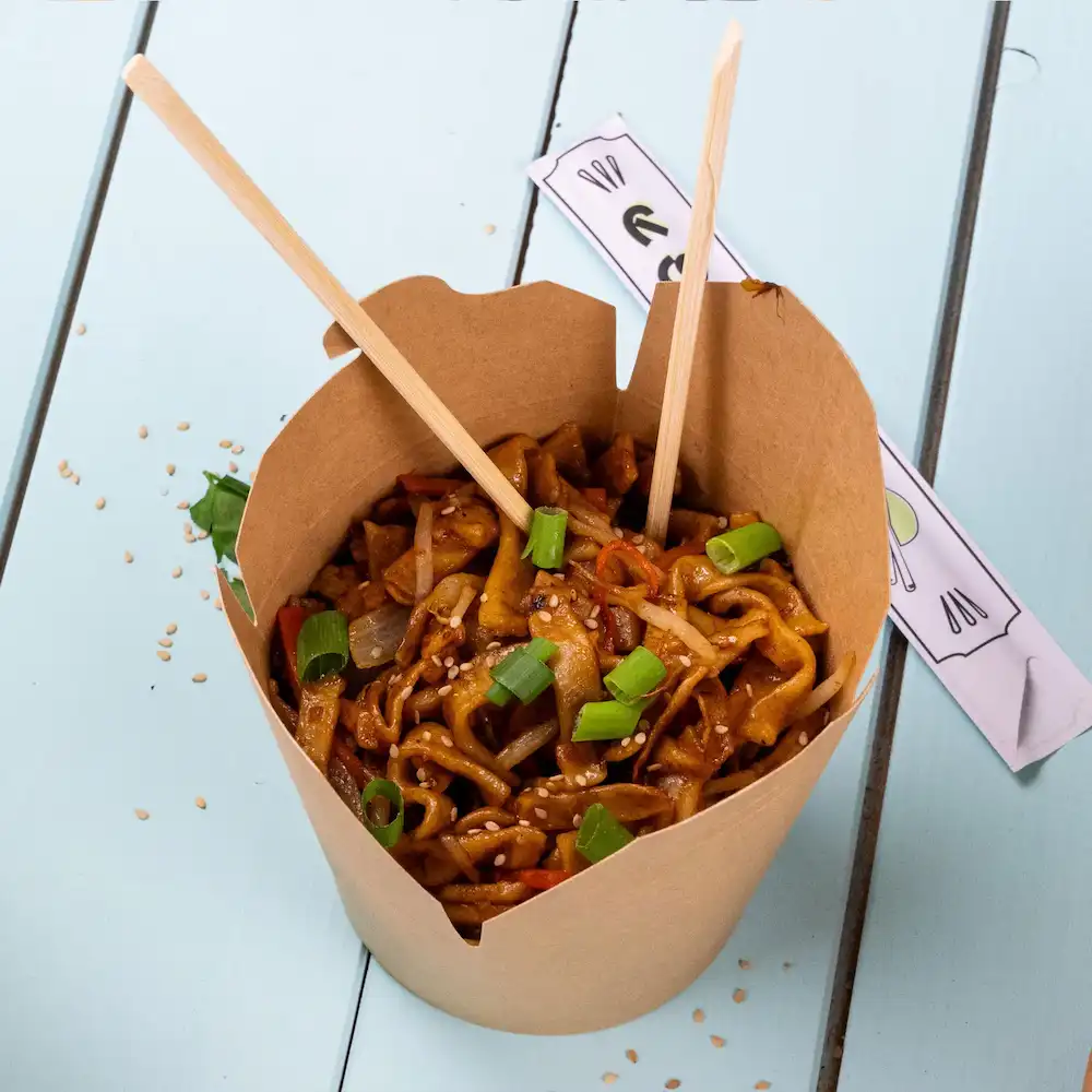 Doos met gebakken noedels met groenten, gegarneerd met lente-ui en sesamzaadjes, geserveerd met chopsticks.