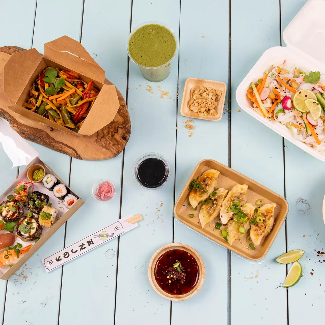 Diverse Aziatische gerechten, waaronder sushi, dumplings, roerbakgroenten en een groene drank op een blauwe houten tafel.