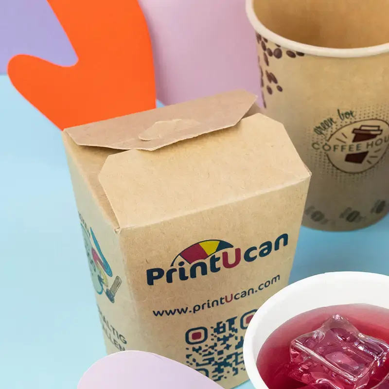 Bruine kartonnen containers voor ramennoedels met een logo, geplaatst op een blauwe ondergrond met verschillende toppings en eetstokjes.