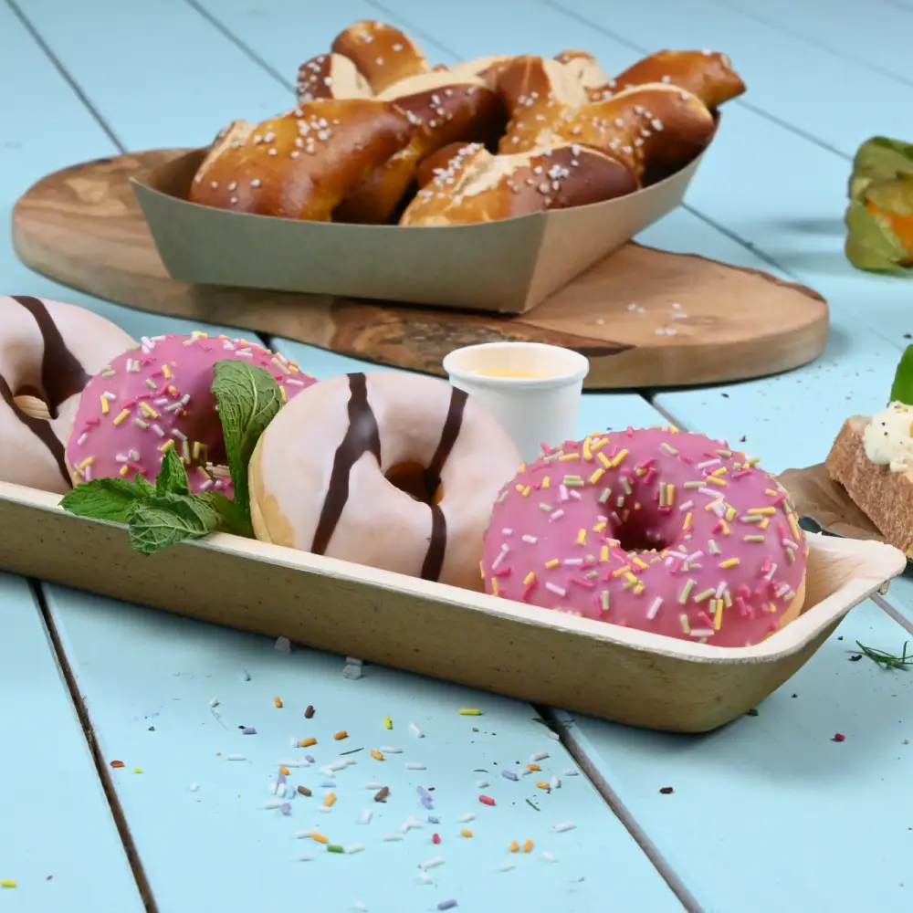 Drie kleurrijke donuts in een papieren tray; twee roze met sprinkles, één met chocoladeglazuur. Pretzels op de achtergrond.