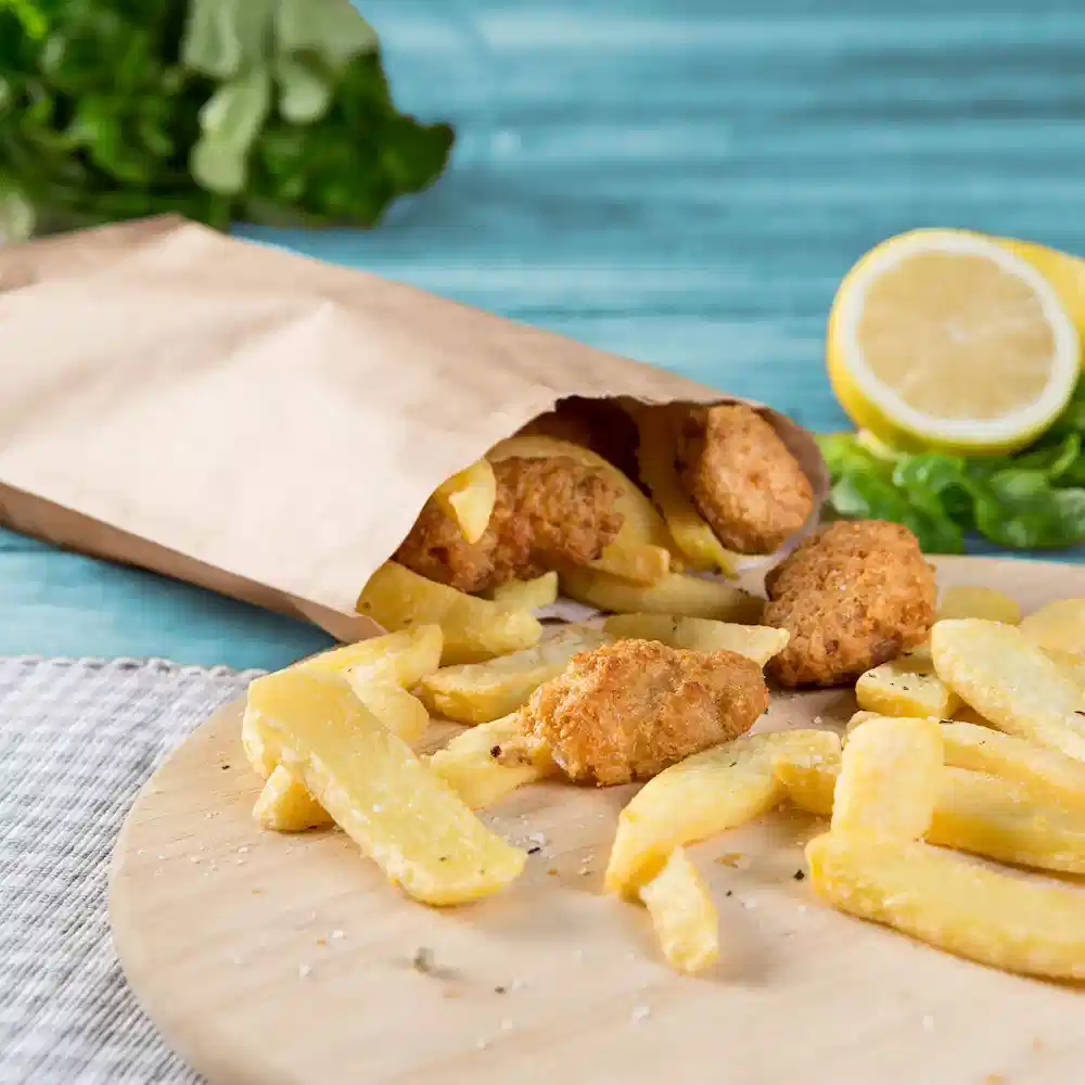 Een papieren zak bevat gefrituurde kipnuggets en gouden frietjes, met een schijfje citroen en groenten op de achtergrond.