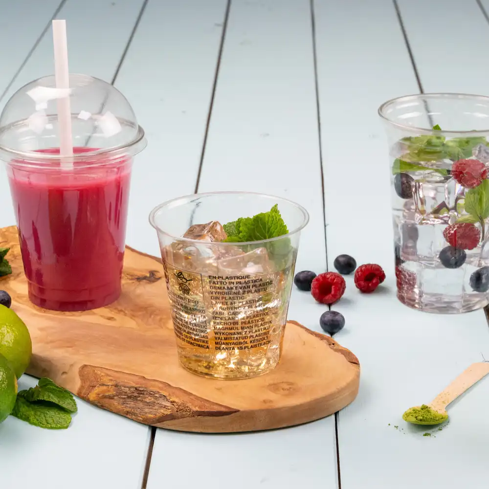 Heldere plastic bekers met drank en munt op een houten plank, met bessen en limoenschelen eromheen.