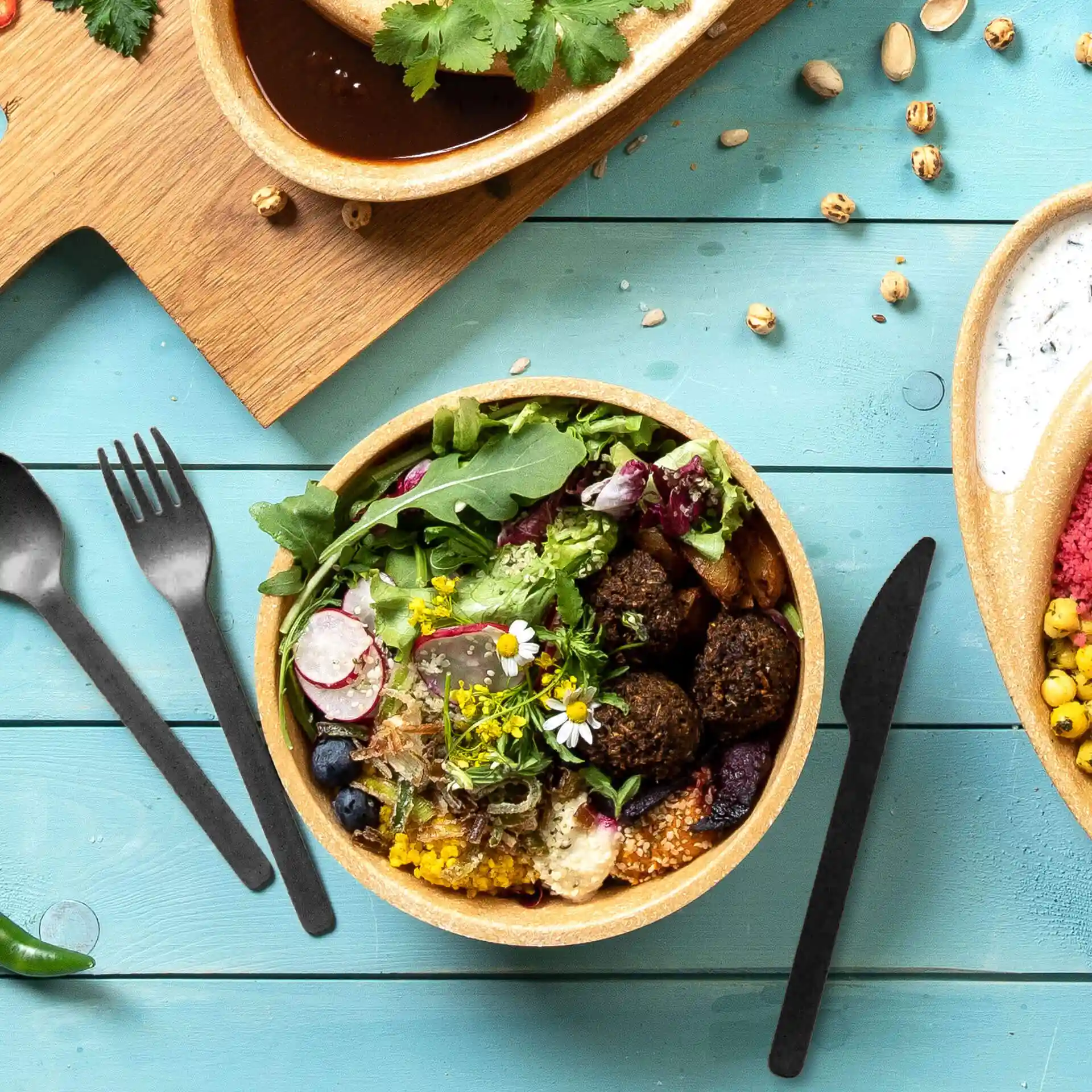 Kom met salade, falafel en eetbare bloemen, vergezeld van zwart herbruikbaar bestek op een blauwe houten ondergrond.