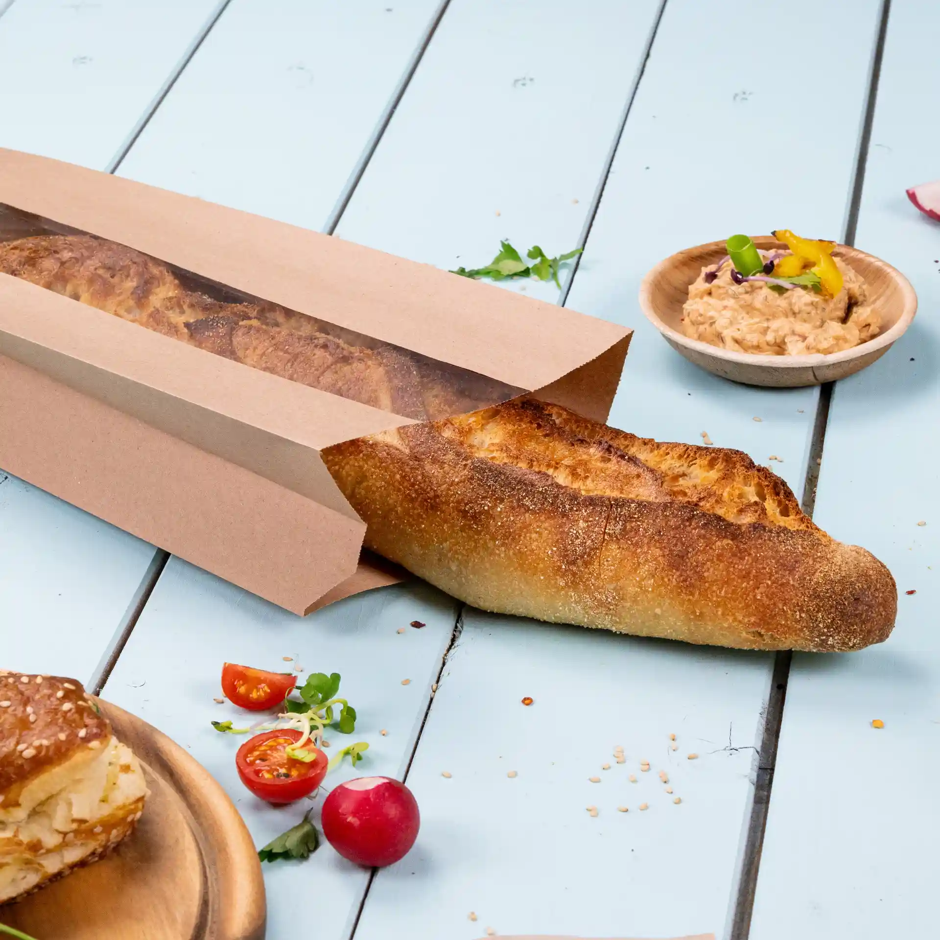 Bruine platte papieren broodzak met een venster, met een baguette erin, op een lichtblauwe houten ondergrond.