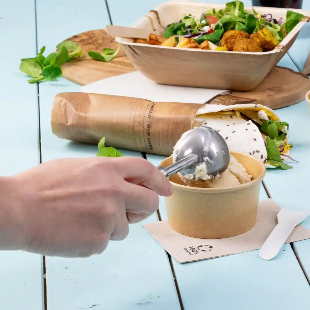 Hand houdt een metalen ijslepel boven een papieren bekertje ijs; salade en taco in milieuvriendelijke containers in de buurt op een blauwe tafel.