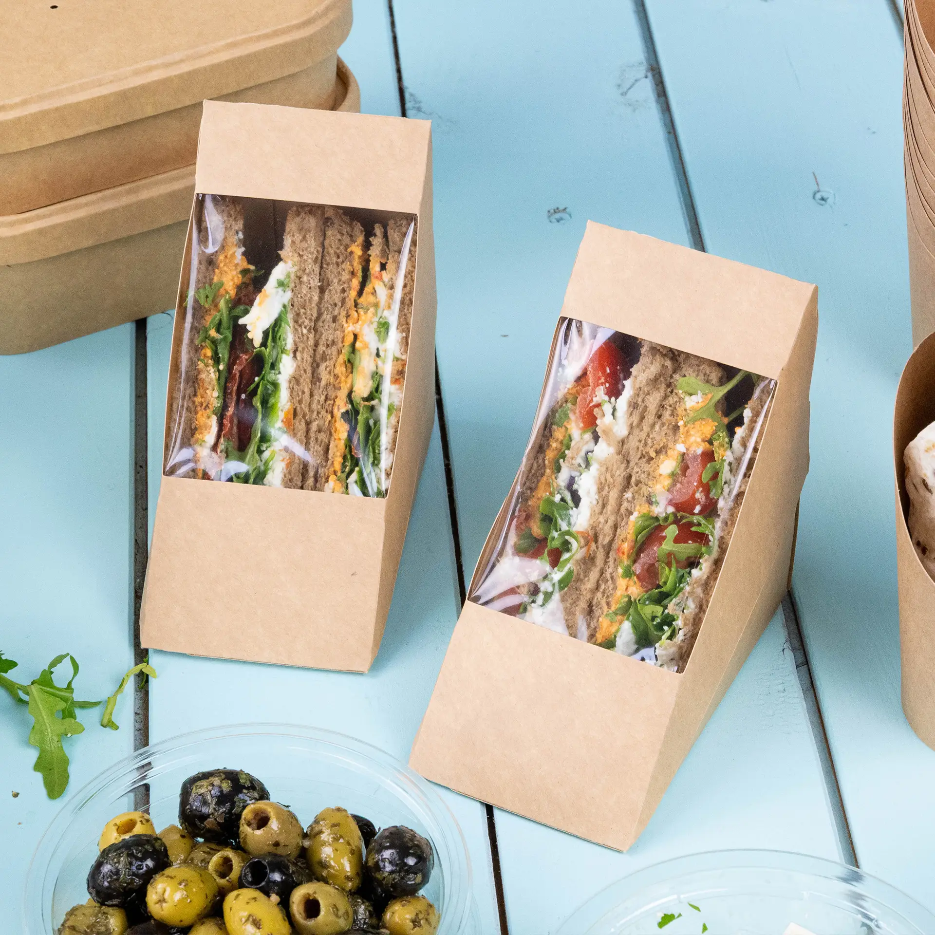 Twee bruine kartonnen sandwichpunten met een doorzichtig PLA-venster dat sandwichs en gevulde broodjes toont op een blauwe ondergrond, met olijven in een kom erbij.