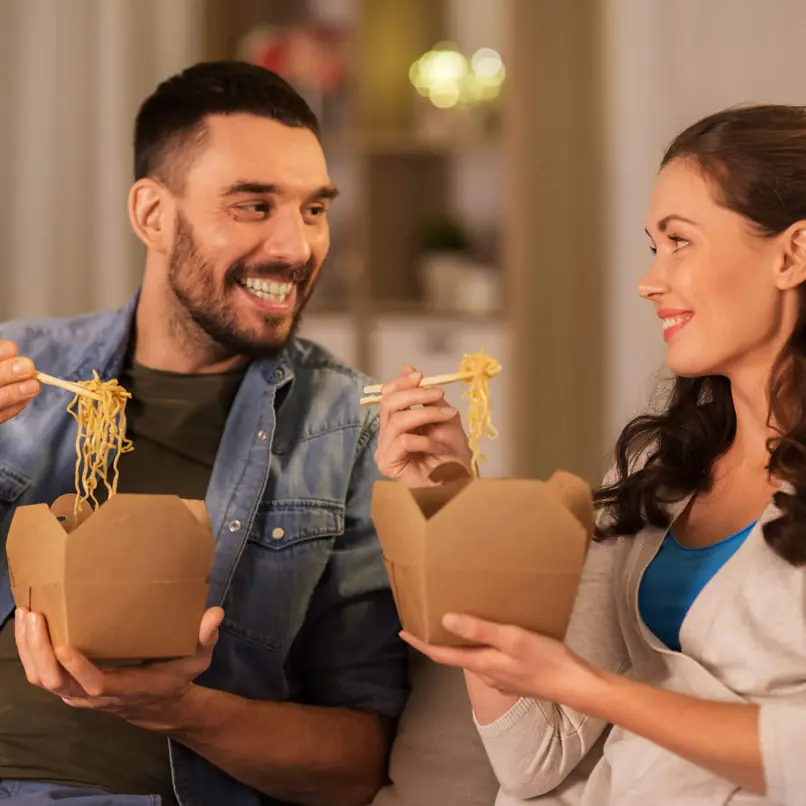 Koppel geniet van afhaalnoedels in bruine dozen, met chopsticks, samen zittend in een gezellige binnenomgeving.