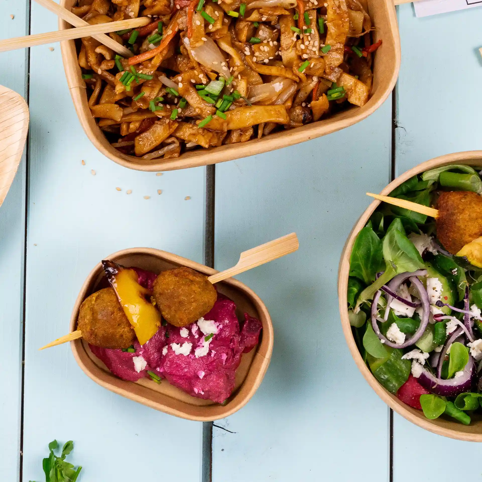 Afbeelding van drie rechthoekige kraft voedselcontainers met verschillende gerechten: noedels, salade en een bietendip met spiesjes.