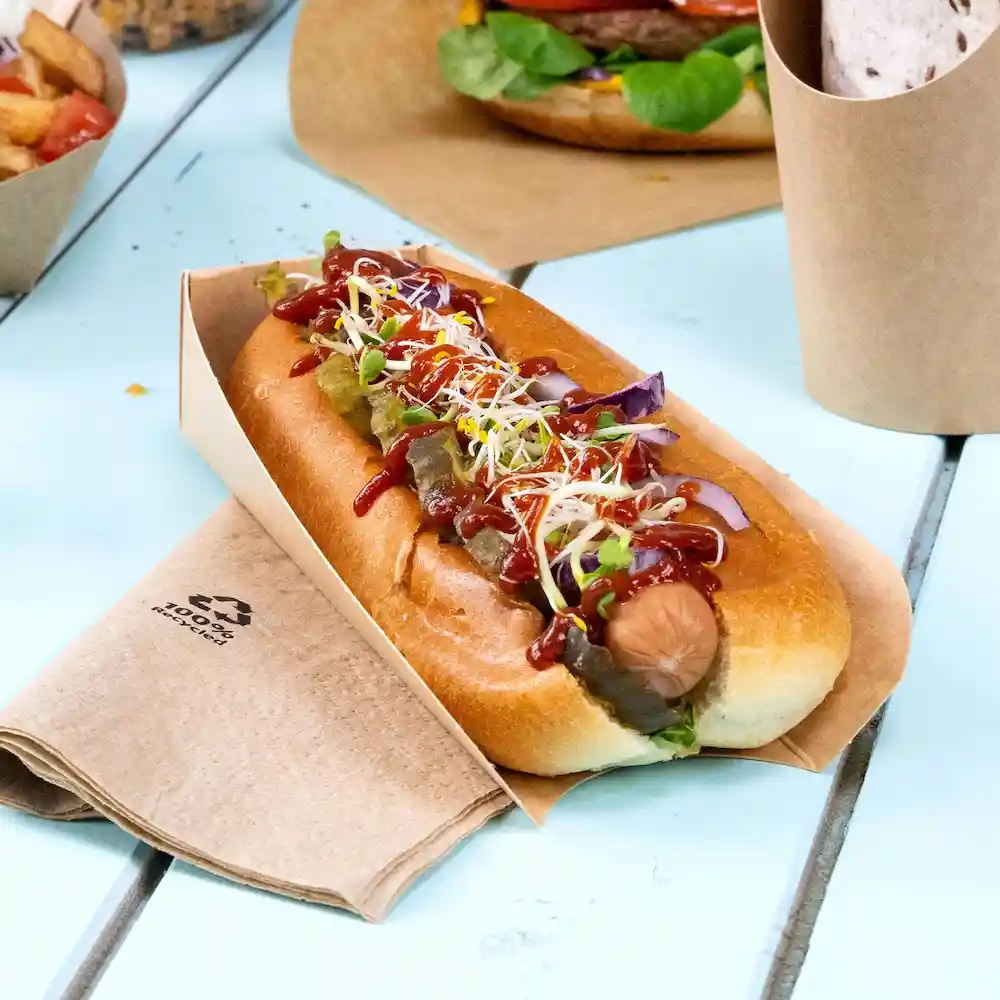 Hotdog in een bruine papieren houder, belegd met ketchup, mosterd, augurken en kiemen, rustend op een houten tafel.