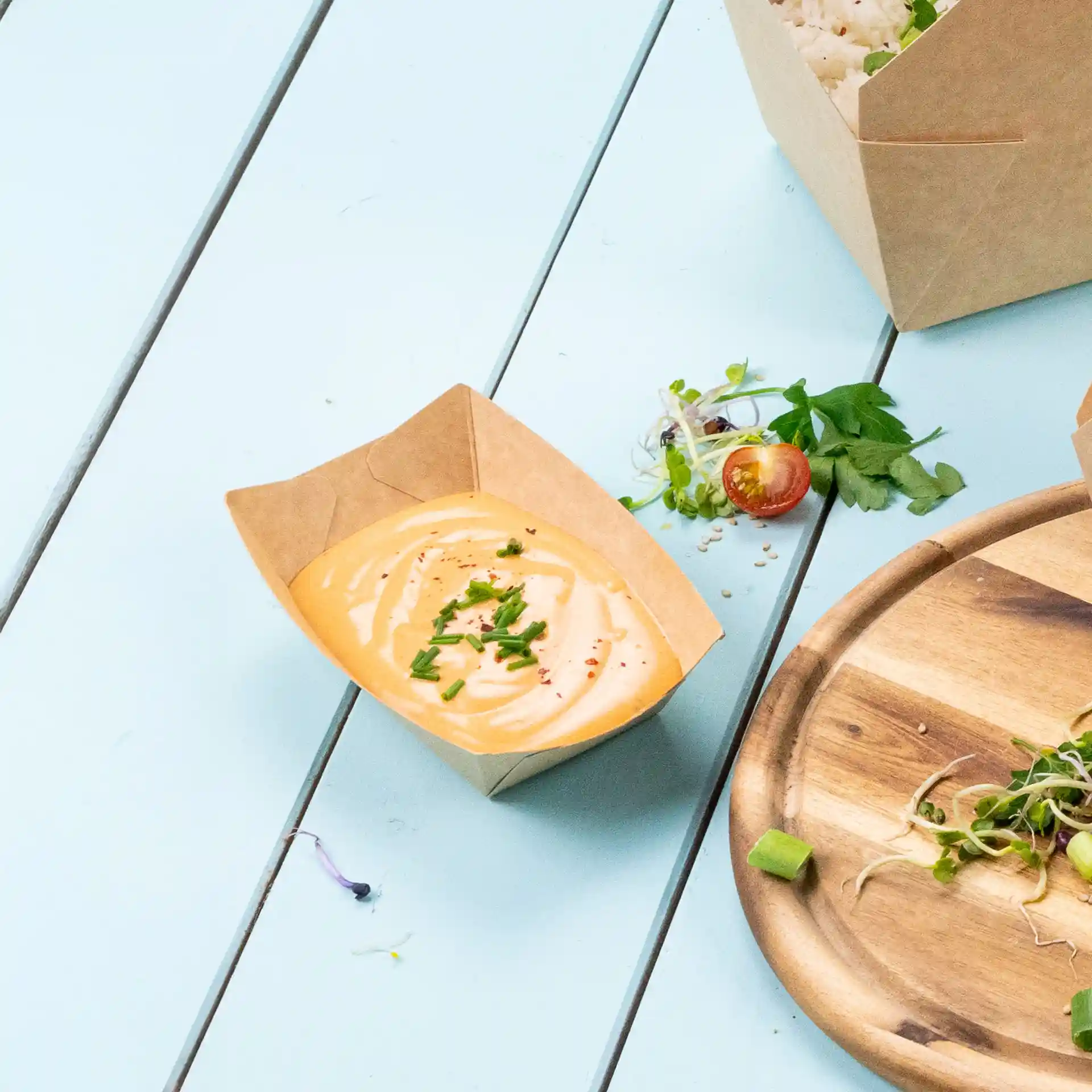 Bruine kartonnen snacktray met een romige saus, getopt met gehakte bieslook, op een lichtblauwe ondergrond.
