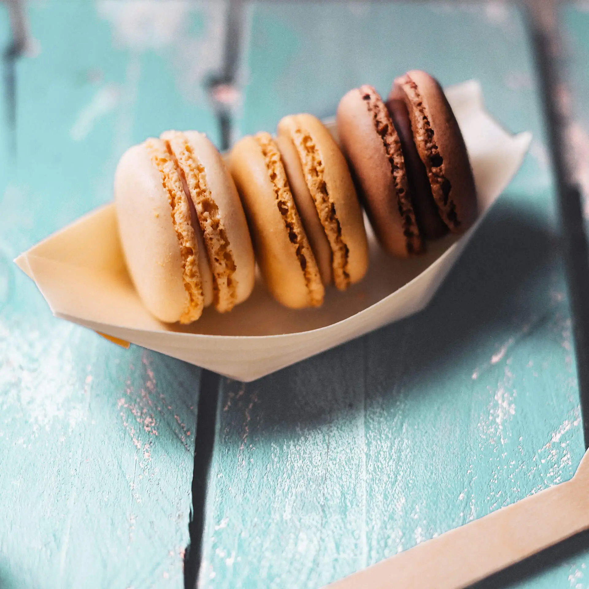 Vier macarons in een bootvormige houder, in beige, lichtbruin, donkerbruin en chocoladekleuren op een gestructureerd oppervlak.