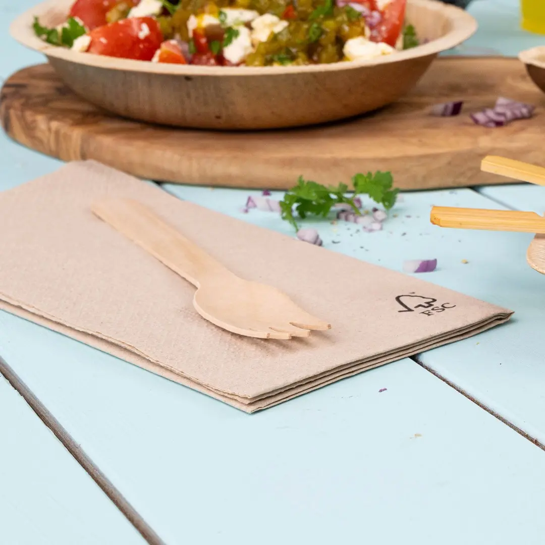 Houten vork op een gevouwen beige servet, met een kom salade op de achtergrond op een lichtblauwe tafel.