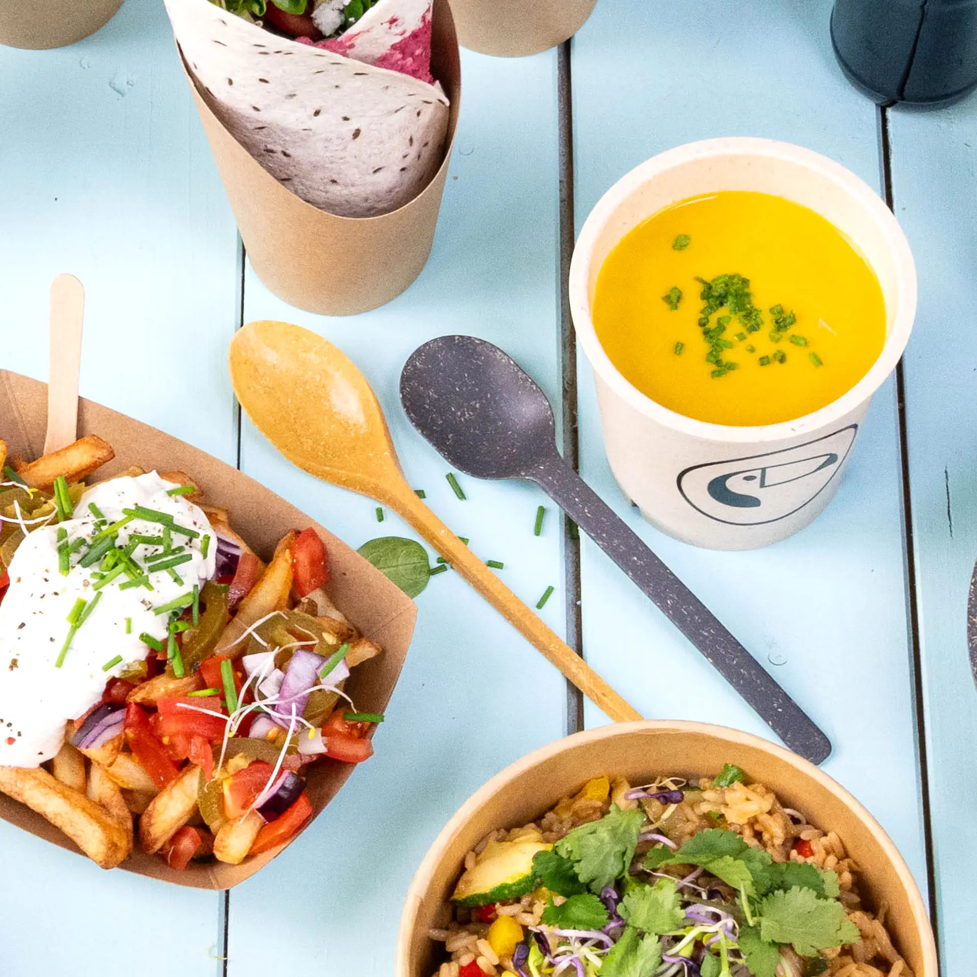 Een bruine herbruikbare lepel van biocomposiet; een kom soep, frieten met toppings en een verpakt item in de buurt.