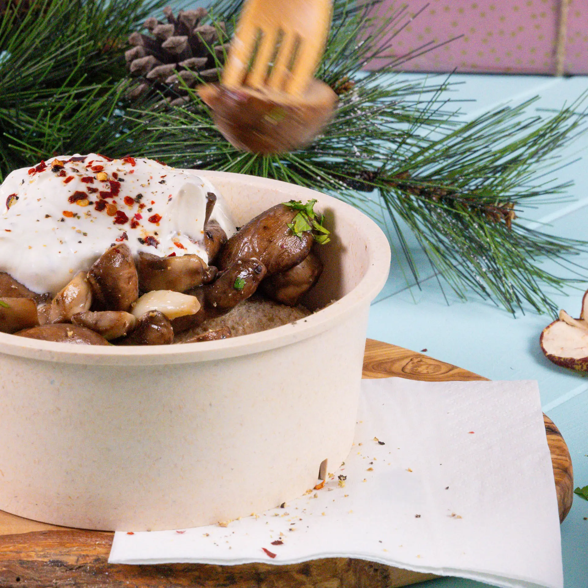 Kom met champignons en yoghurt, getopt met rode pepervlokken, op een houten plank met een witte servet ernaast.
