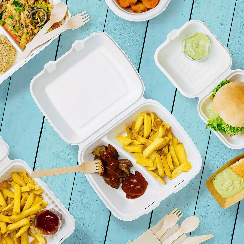 Witte takeaway-containers met friet, worst en sauzen op een blauwe houten ondergrond, naast een burger en andere etenswaren.