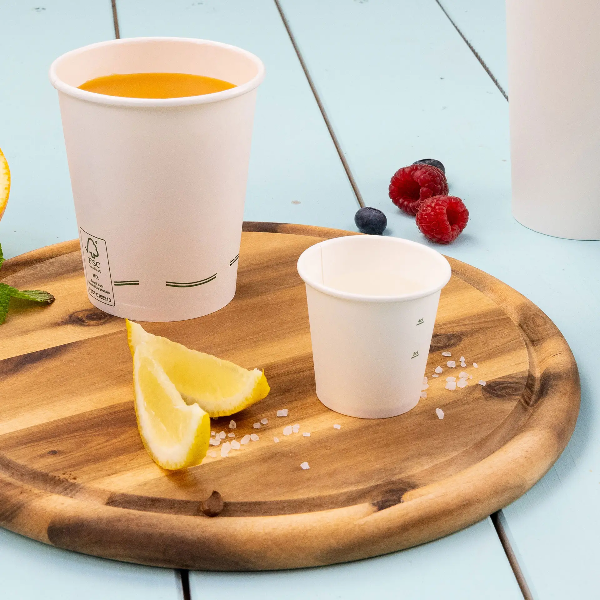 Een witte papieren bekertje van 2 oz op een houten schaal, met een grotere beker gevuld met sinaasdrank, citroenpartjes en bessen in de buurt.