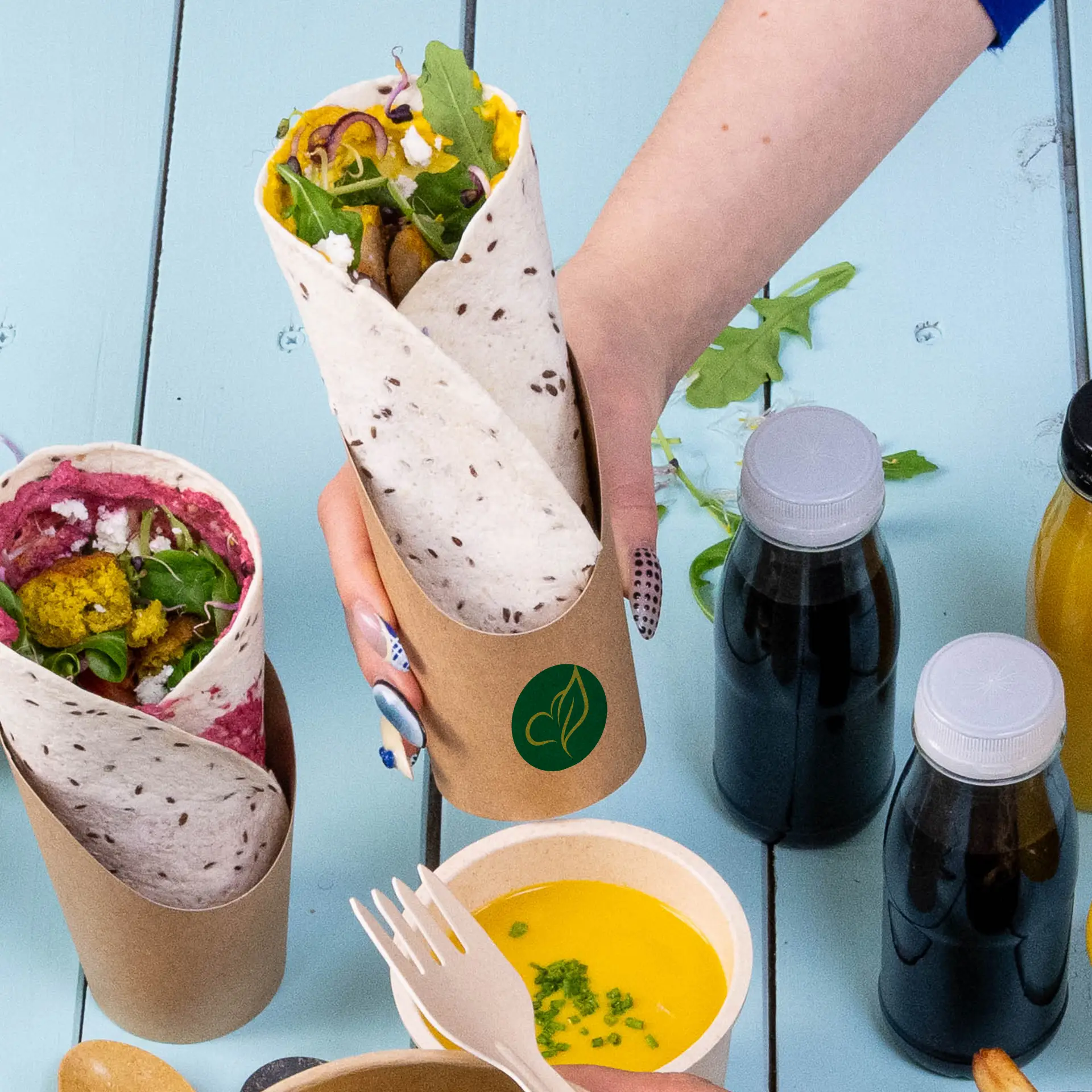 Een kraft kartonnen wrapbekertje met een gevulde wrap; een ander bekertje met een kleurrijke wrap ernaast, omringd door dranken en een dipsaus.