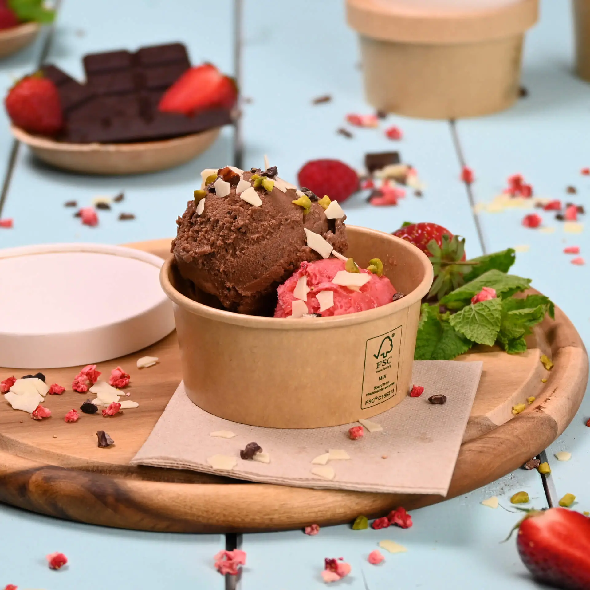 Bruine papieren ijscoupe met chocolade- en aardbeienijs, getopt met noten en omringd door verse aardbeien en munt.