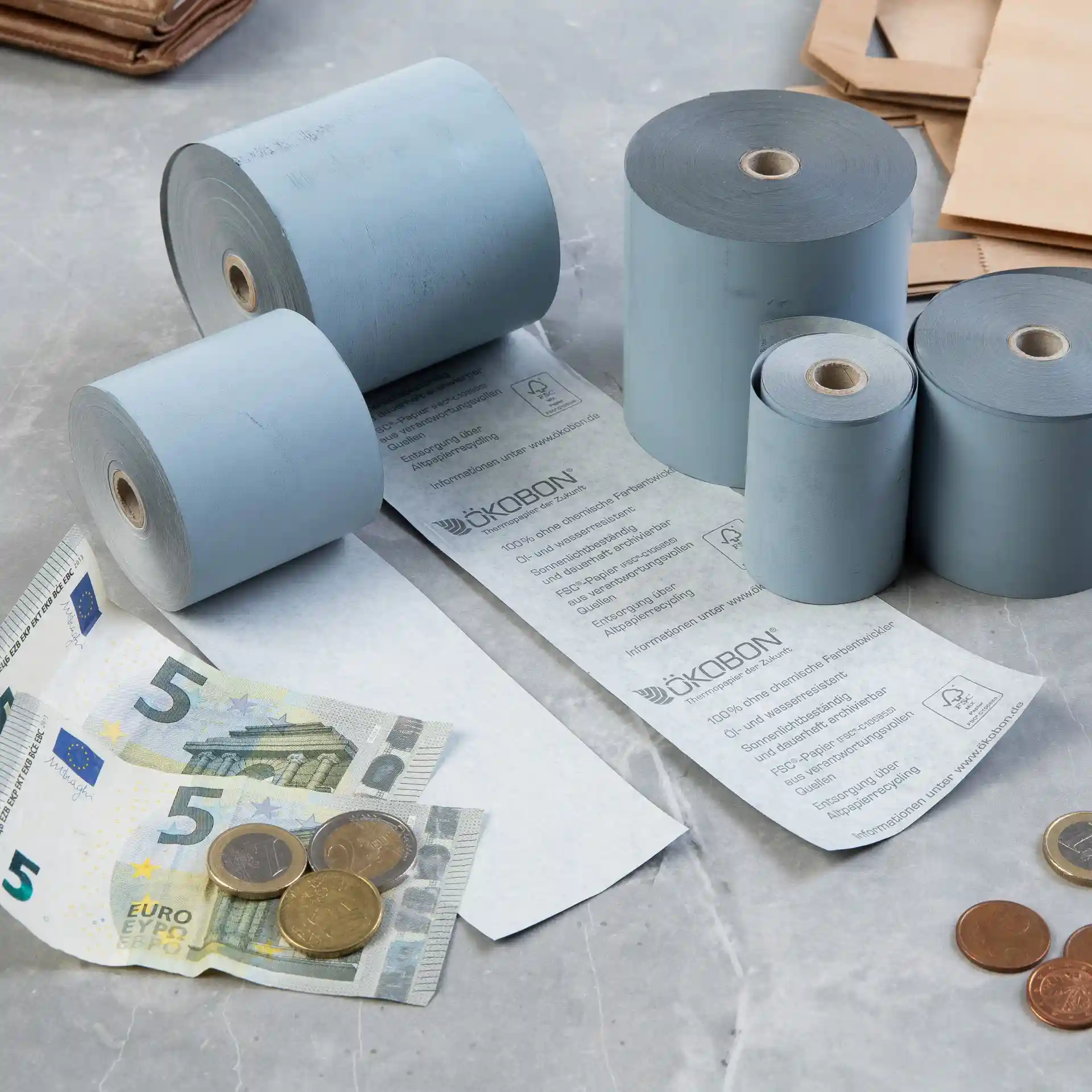 Afbeelding van meerdere grijze thermopapierrollen, een 5-eurobiljet en munten op een oppervlak.