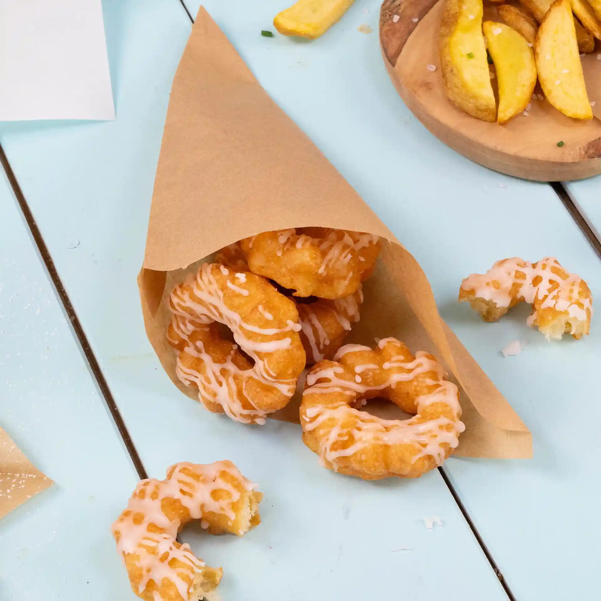 Bruine papieren zak met meerdere gefrituurde donuts met glazuur, geplaatst op een lichtblauwe ondergrond.