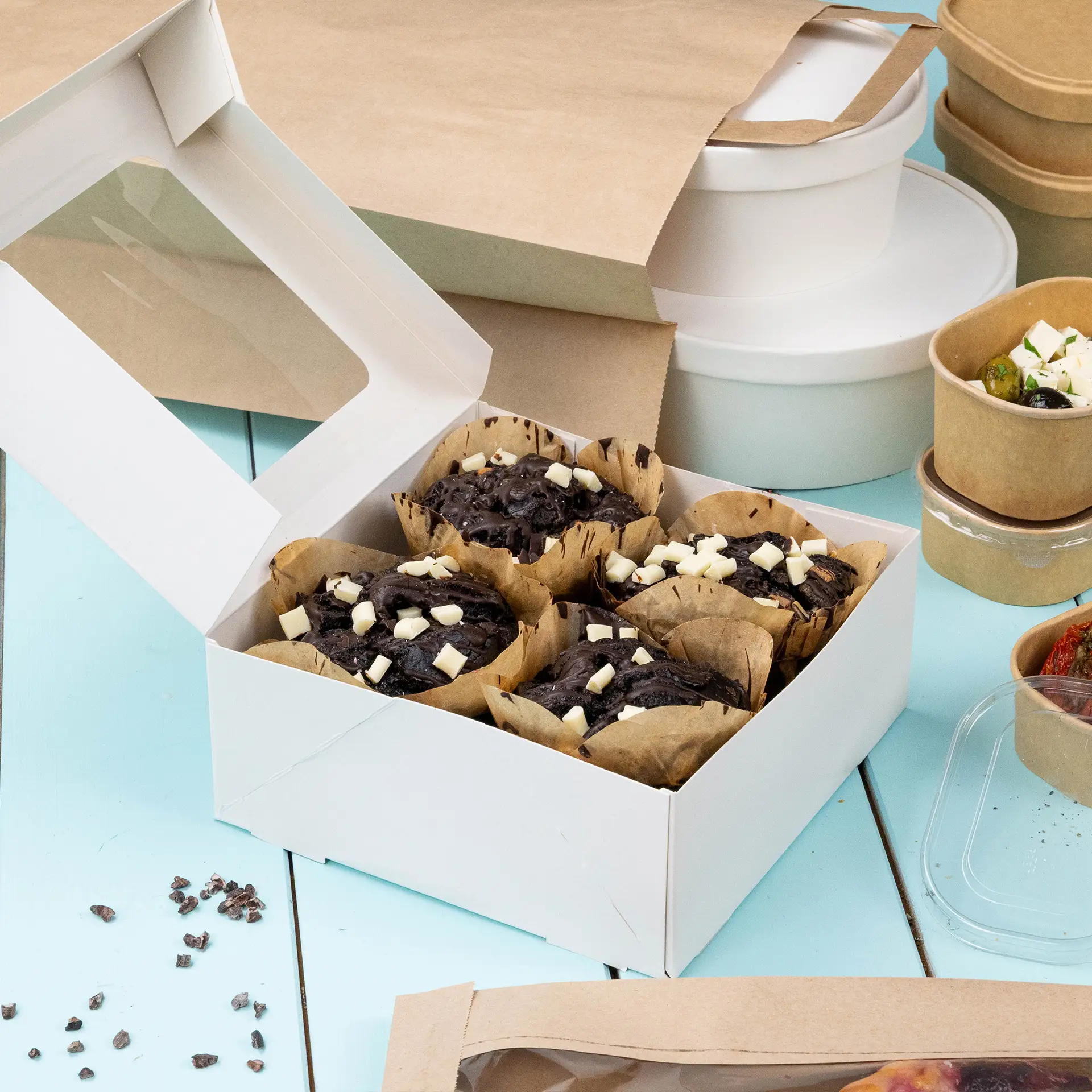 Een witte patisseriebox met een cellulosevenster met chocoladegebak; diverse andere verpakkingsitems in de buurt.