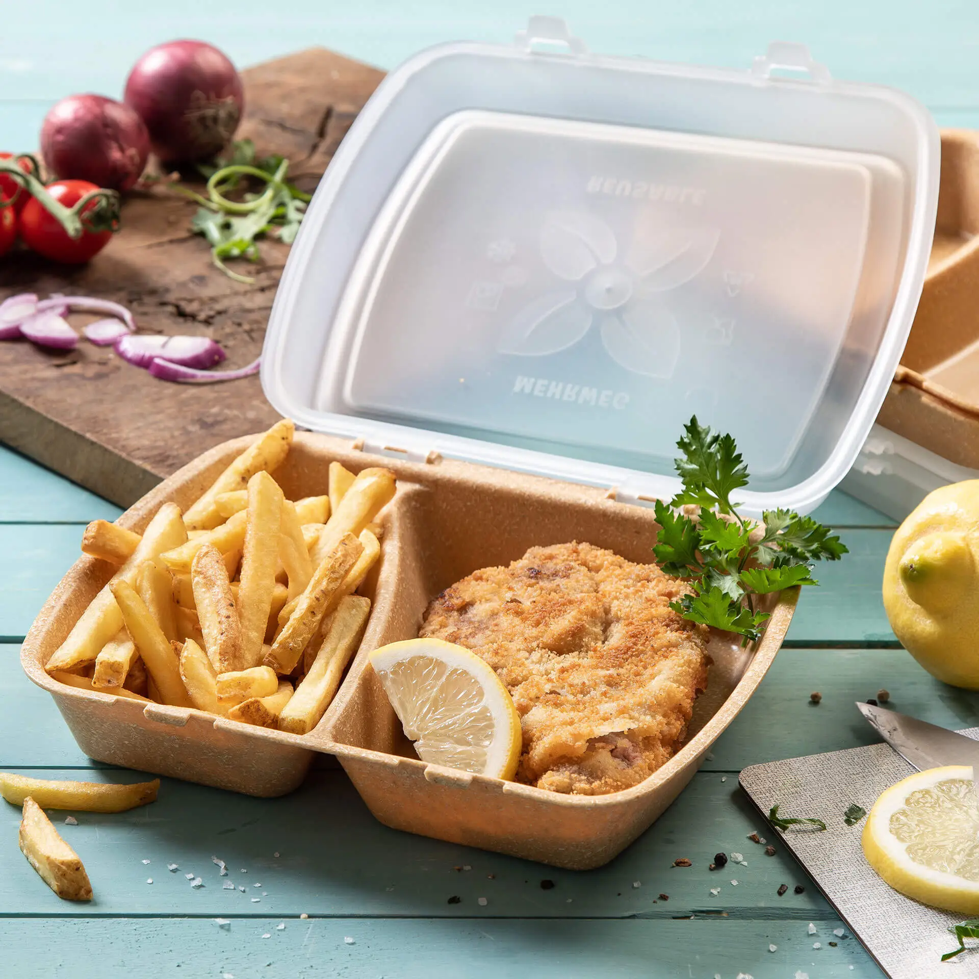 Container met gefrituurde kip, frietjes en een schijfje citroen in een doorzichtige plastic doos op een blauwe ondergrond.