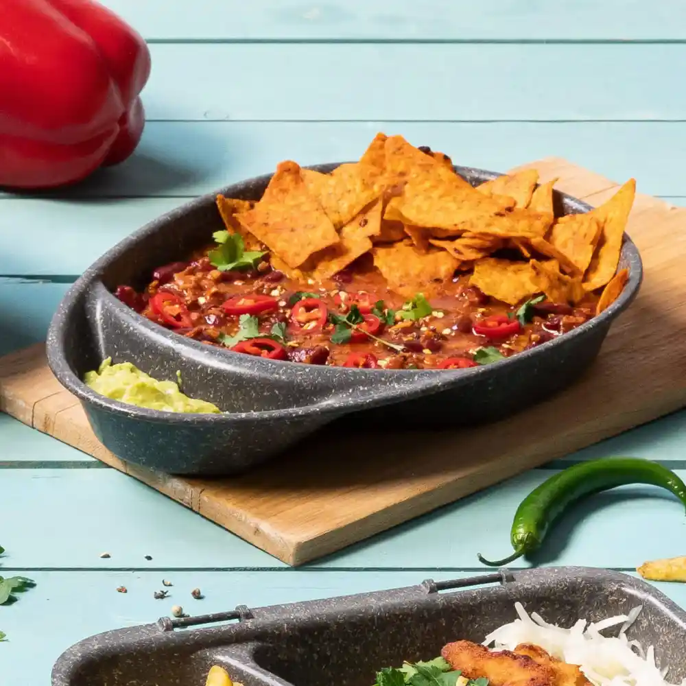 Kom met chili, belegd met tortillachips, gesneden rode chili's en koriander op een houten plank met toppings.