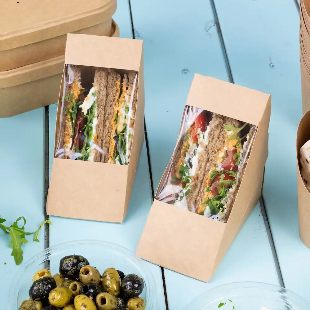 Twee sandwichdozen van bruin kraftpapier met vensters van gelaagde sandwiches en een schaal met gemengde olijven.