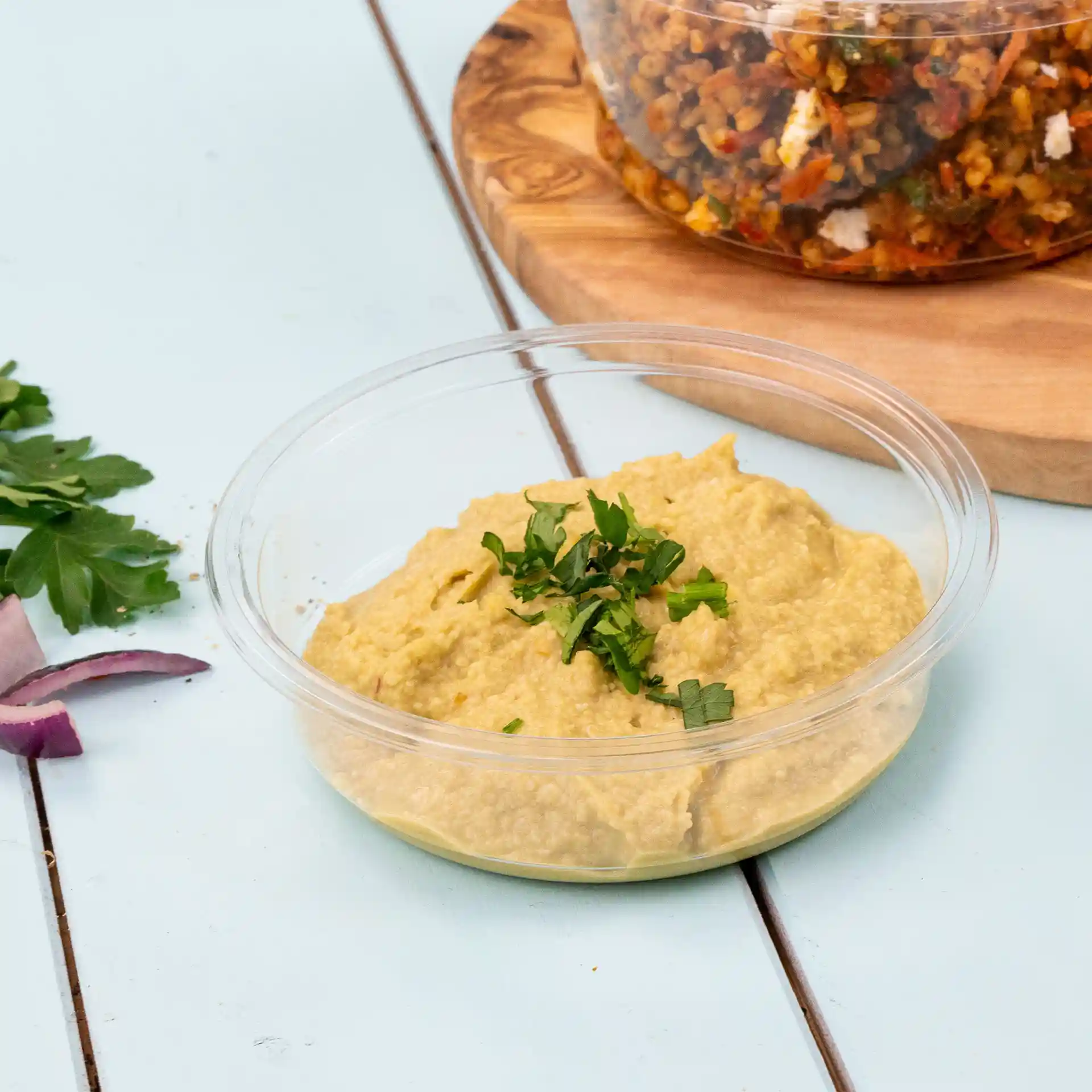 Ronde transparante deli-container met hummus, getopt met gehakte peterselie, op een lichtblauwe ondergrond.