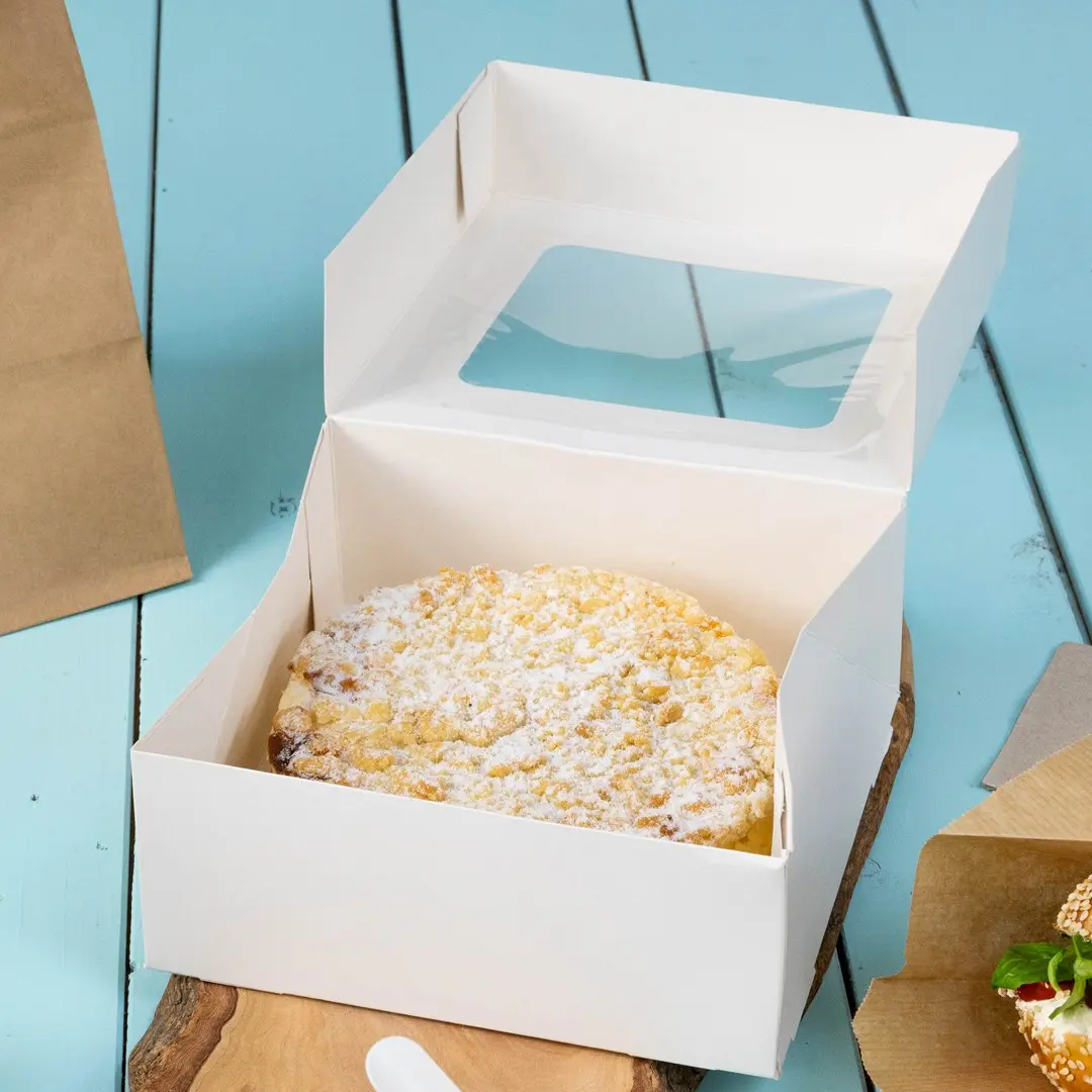 Witte cakebox met een helder venster, bevat een ronde cake bestrooid met poedersuiker, op een houten oppervlak.