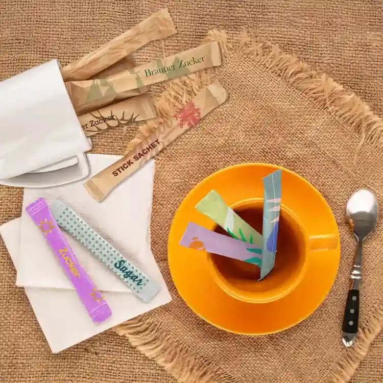 Assortiment van suikerstick zakjes in een beker thee op een juten oppervlak, met een lepel en servet erbij.