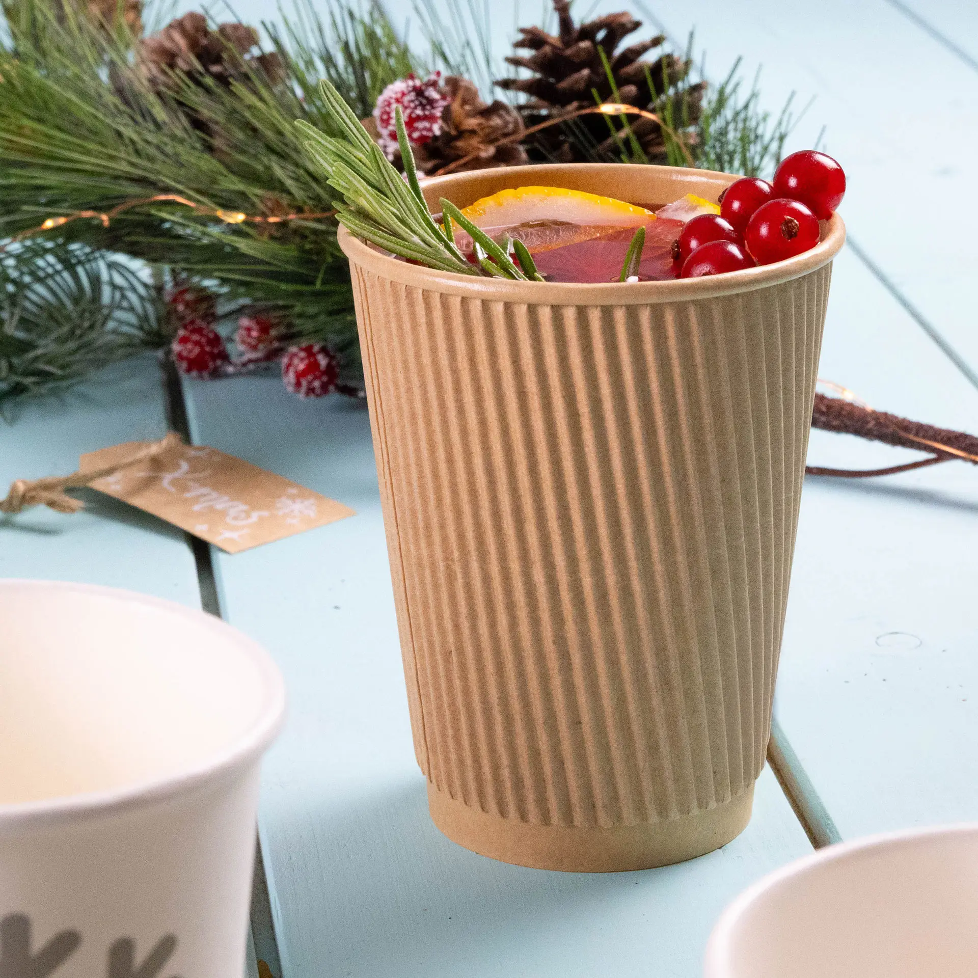 Een kraftbruine 12 oz ribbelbeker gevuld met een warme drank, gegarneerd met sinaasappelschijfjes en bessen, op een lichtblauwe ondergrond.