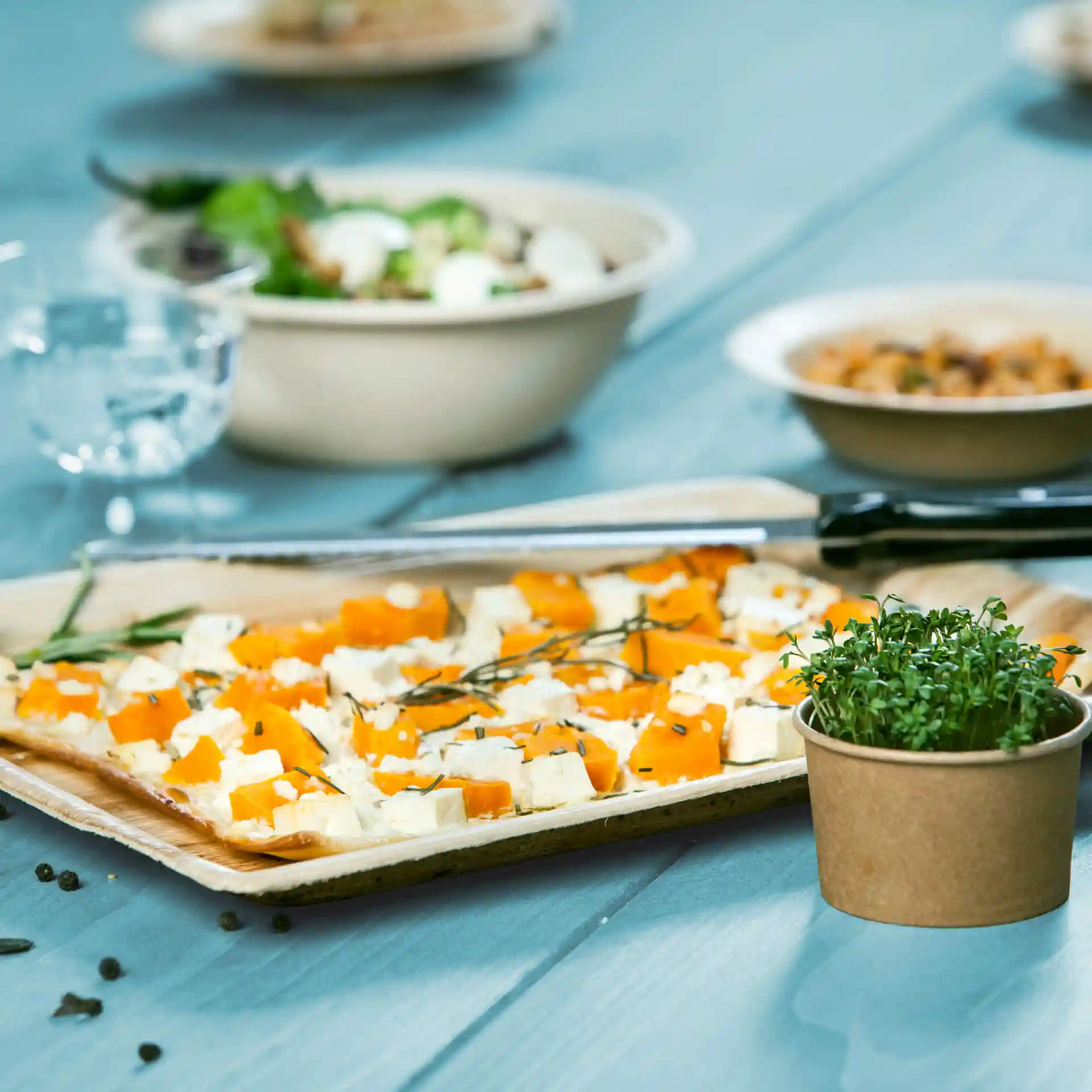 Rechthoekig palmblad dienblad met oranje en witte voedselstukken, vergezeld van een klein potje microgroenten op een blauwe tafel.