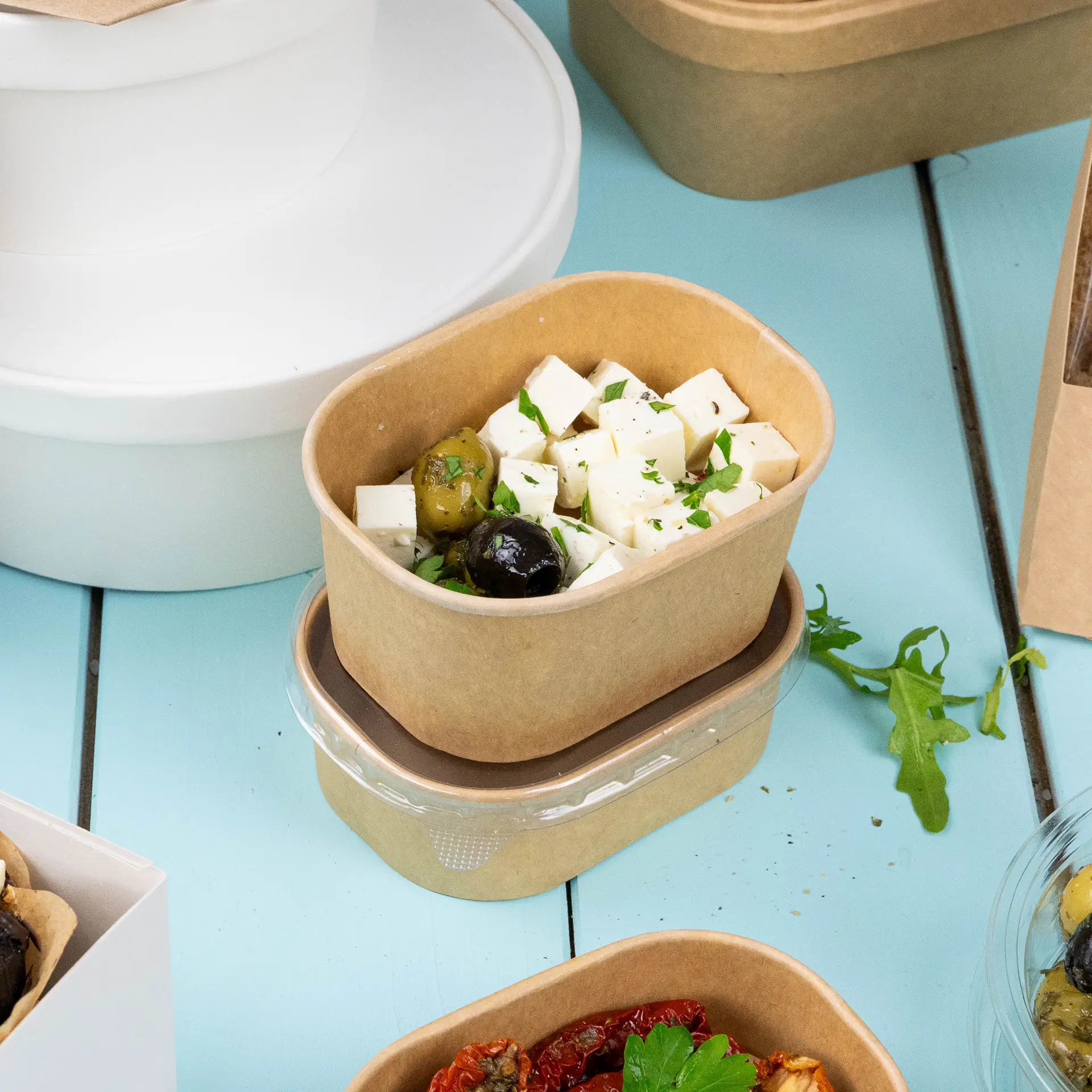 Een kraft voedselcontainer gevuld met blokjes feta kaas en olijven, met een andere vergelijkbare container eronder.