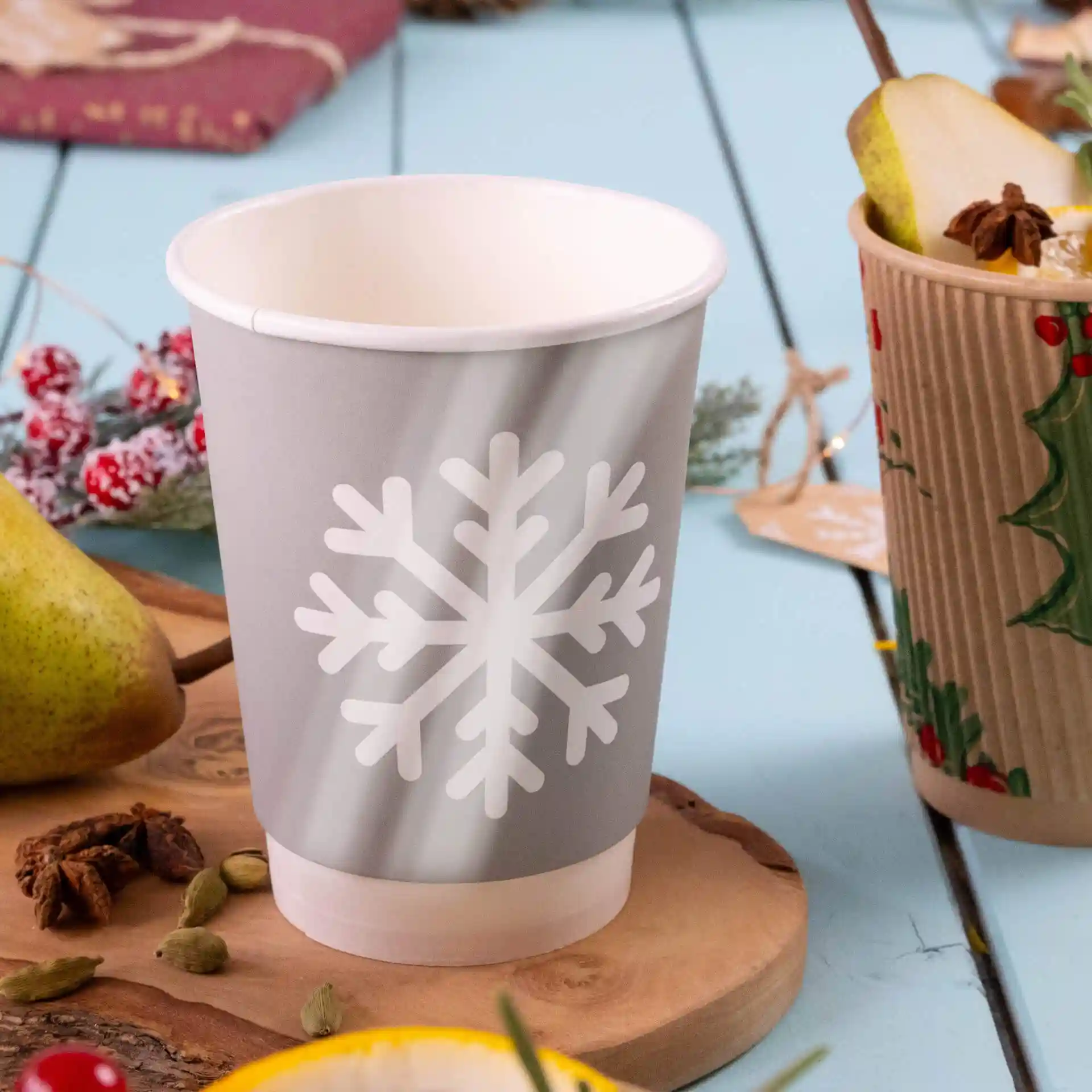 Grijze papieren beker met een wit sneeuwvlokdesign, dubbelwandig, op een houten oppervlak met een peer en specerijen erbij.