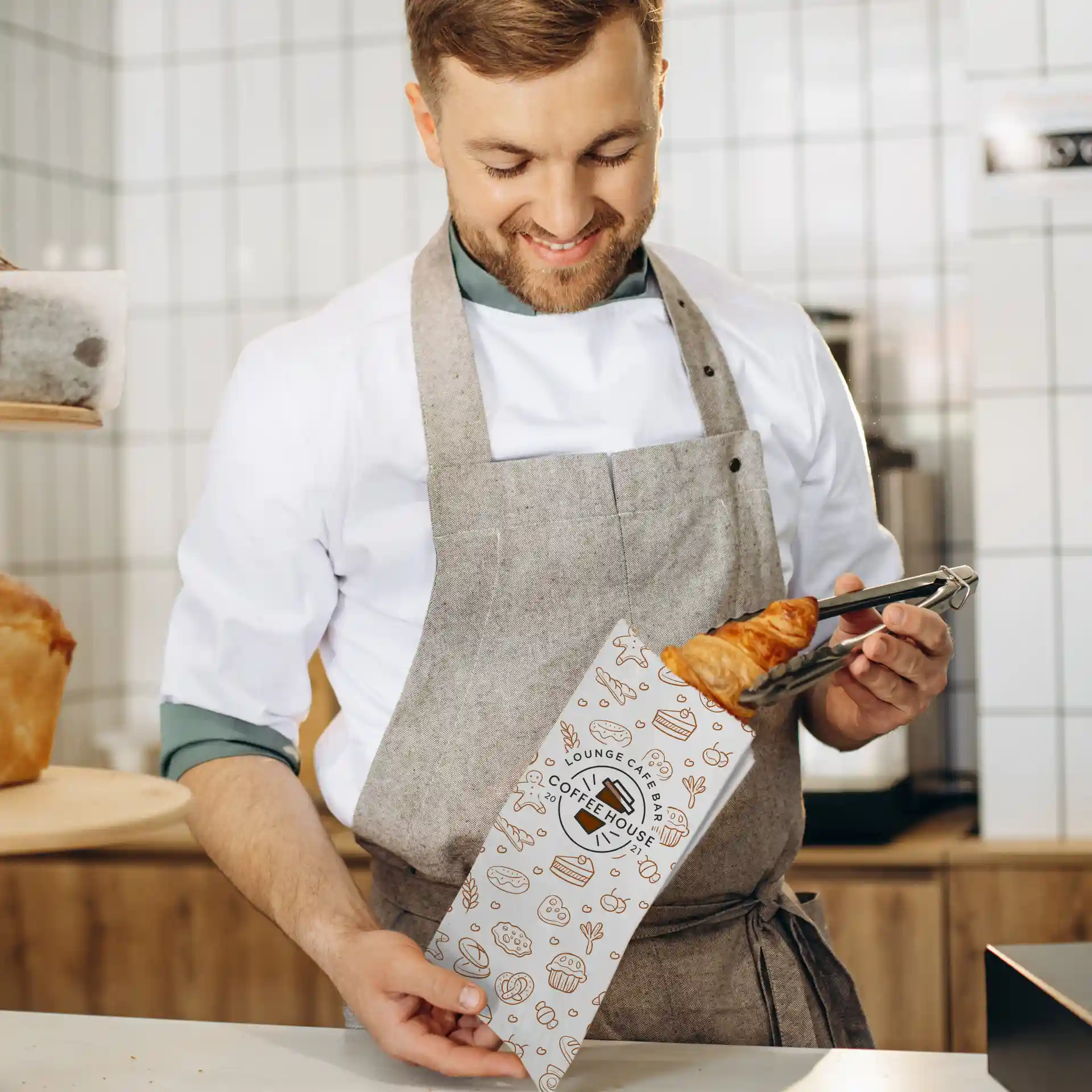 Een persoon in een schort houdt een bedrukte kartonnen broodzak met een croissant in een café.