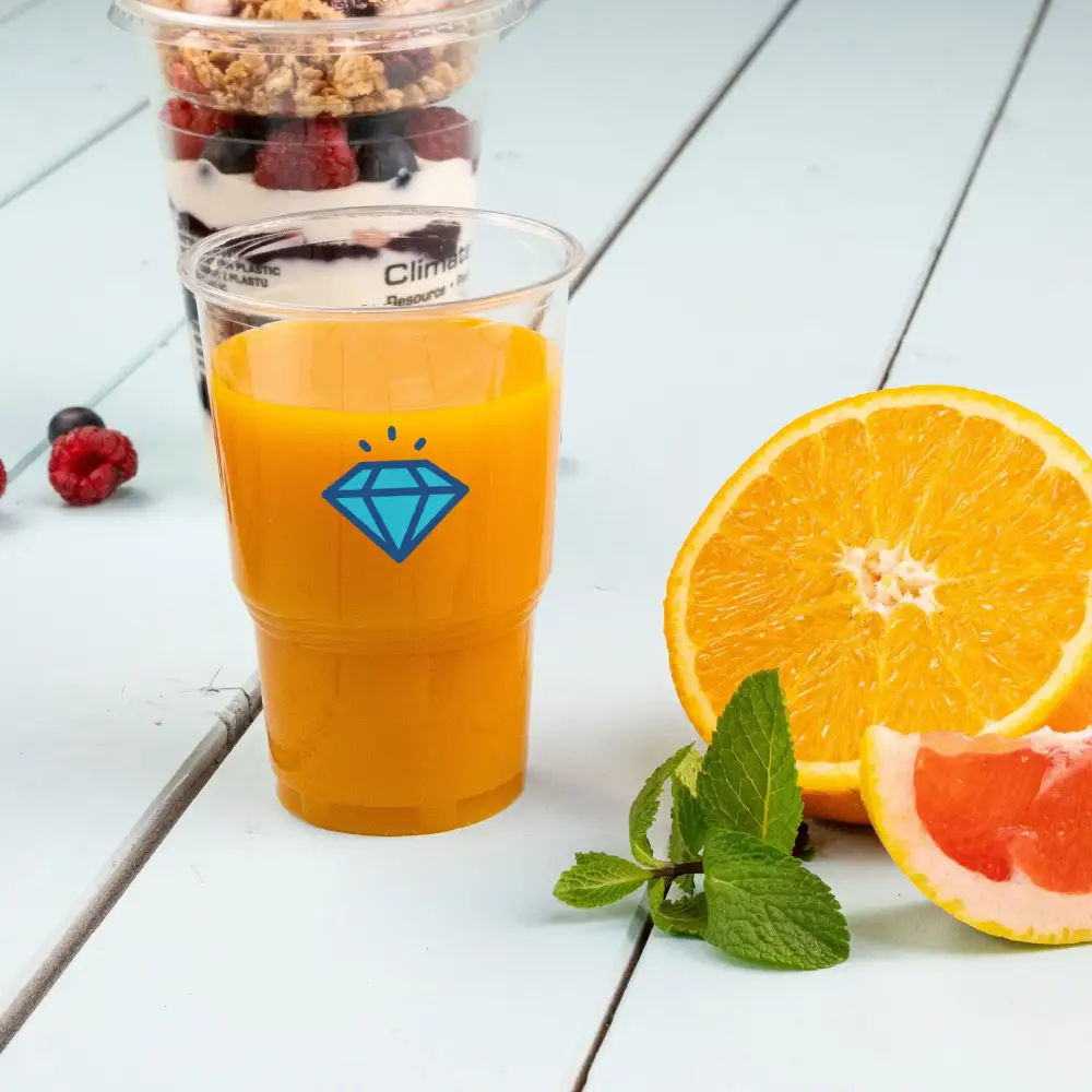 Heldere plastic beker met sinaasappelsap en een blauw diamantpatroon, vergezeld van een gesneden sinaasappel en muntblaadjes.