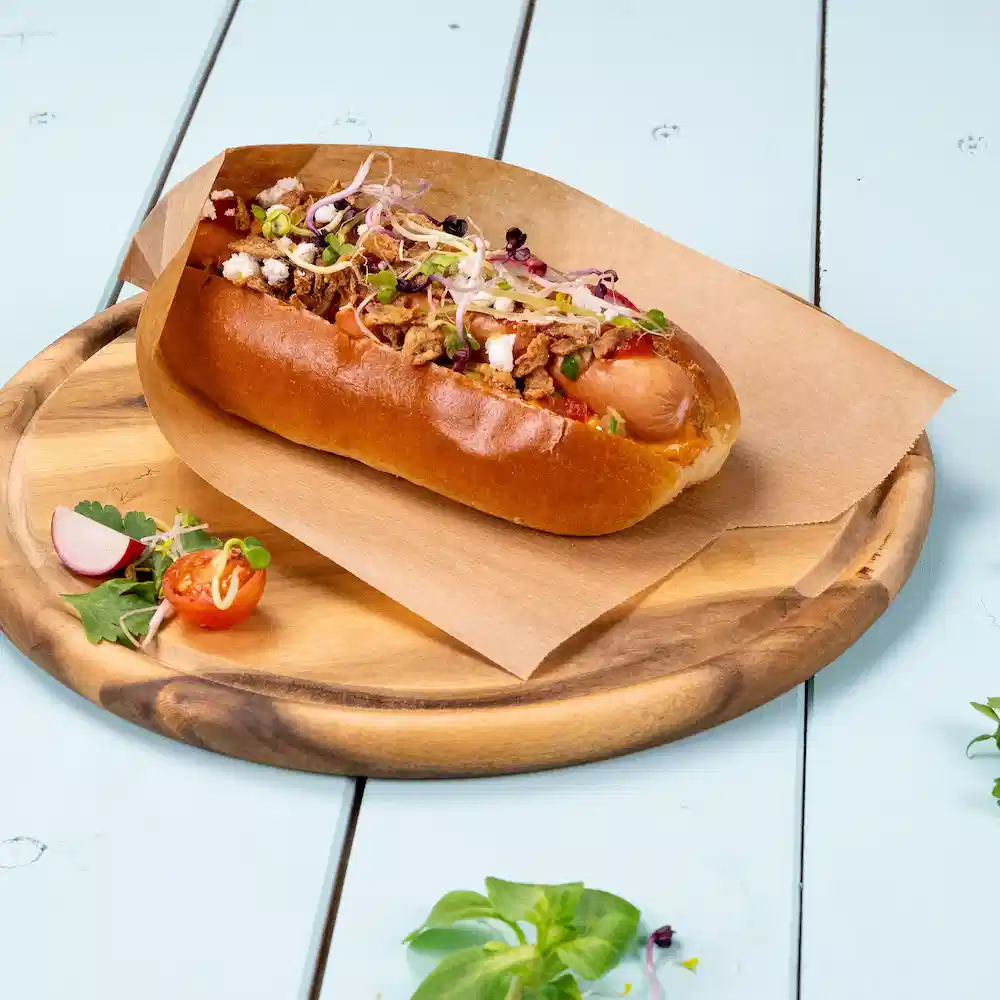 Hotdog in een broodje, belegd met uien, kruiden en knapperige toppings op een houten schaal met groenten erbij.