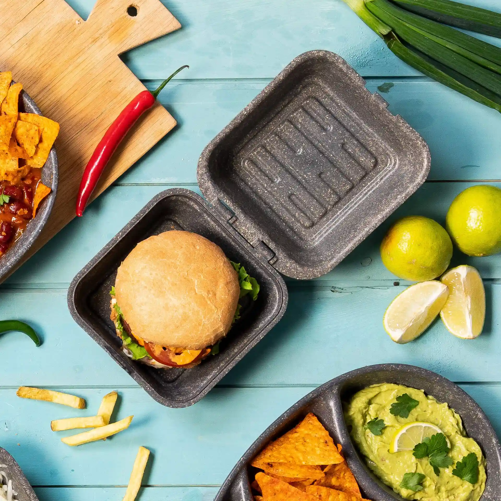Herbruikbare grijze burgerdoos met deksel, bevat een burger, omringd door limoen, nacho's en andere etenswaren.