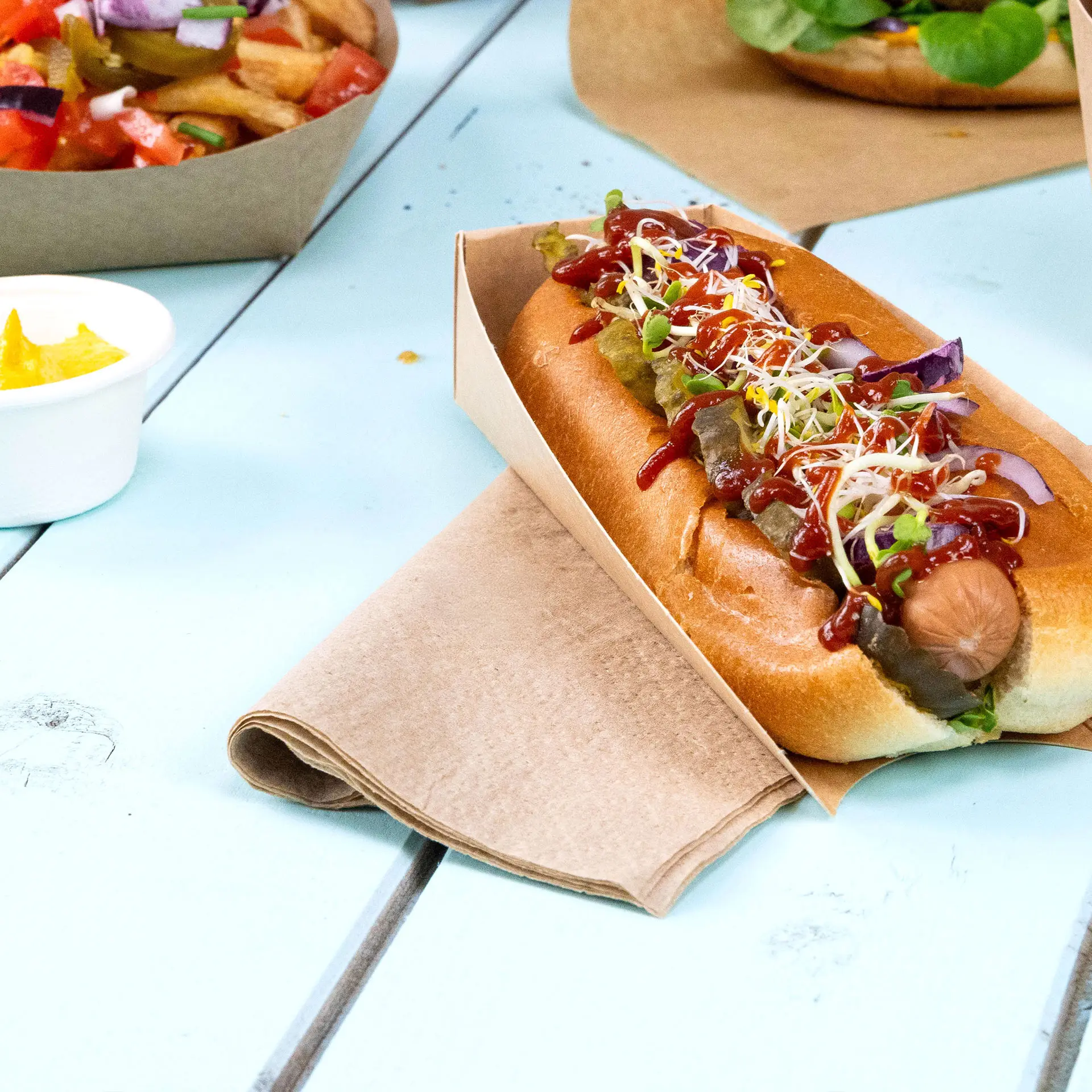Hotdog in een papieren houder, belegd met ketchup, mosterd, augurken en kiemen, met een servet ernaast op een blauwe tafel.