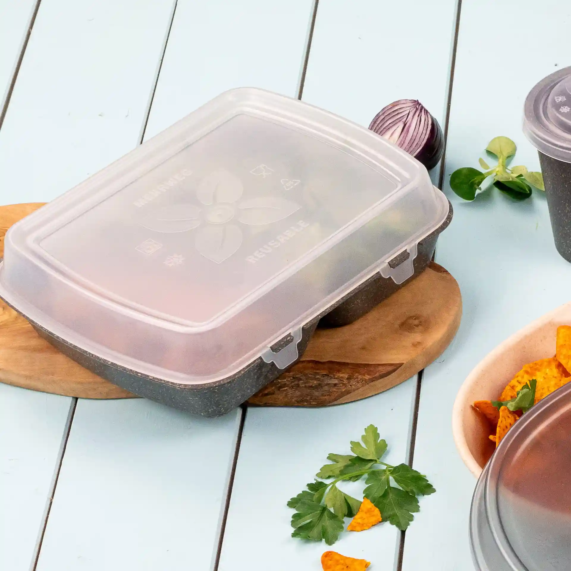 Transparante herbruikbare voedselcontainer met deksel, op een houten plank, omringd door kruiden en snacks.