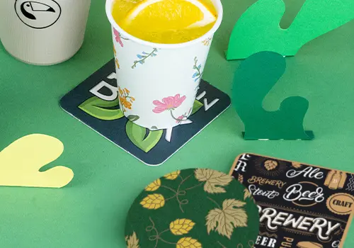 Stapels onderzetters op een houten oppervlak; sommige hebben een logo met een vogel, andere een ontwerp met bier en tekst.