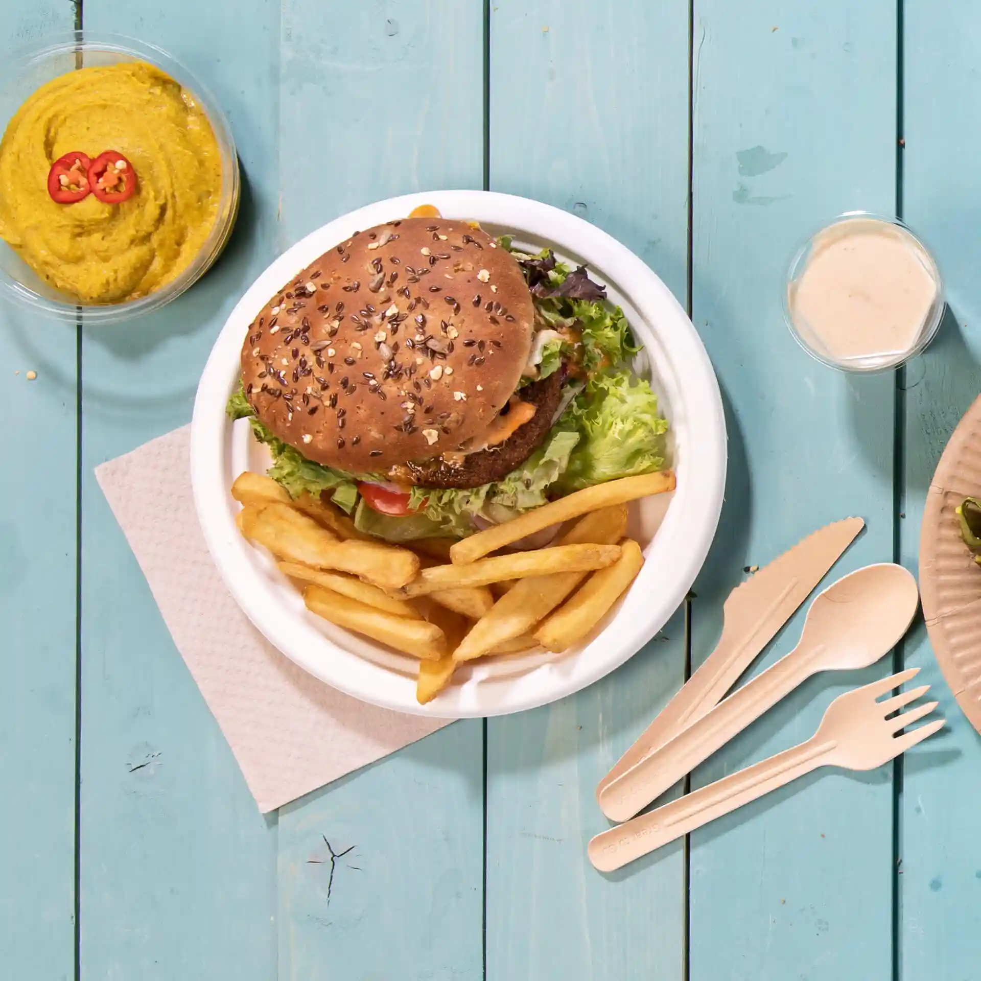 Afbeelding van een rond wit suikerriet bord met een hamburger, frietjes en een kom hummus op een blauwe houten ondergrond.