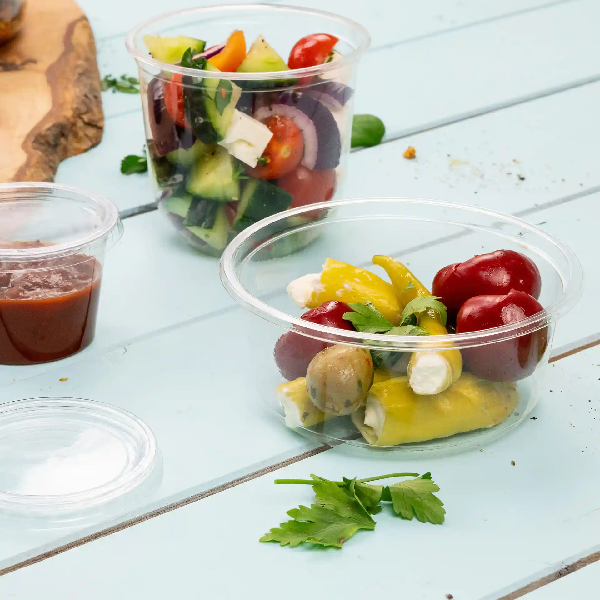 Transparante ronde deli-container met verschillende groenten, olijven en kruiden op een blauwe houten ondergrond.