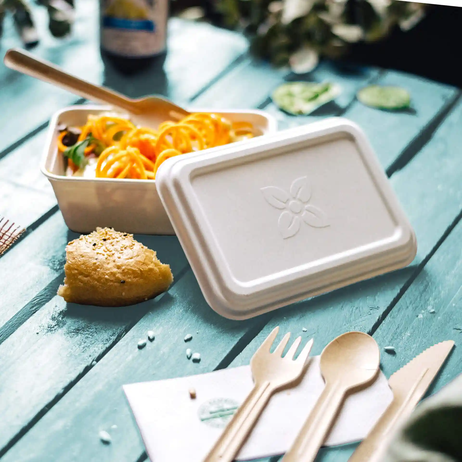 Witte rechthoekige suikerbietencontainer met deksel, vork, lepel en een stuk brood op een blauwe houten ondergrond.