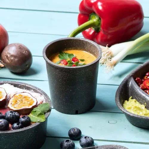 Herbruikbare soepbeker gevuld met soep en gegarneerd met kruiden, omringd door verse groenten en fruit op een blauwe ondergrond.